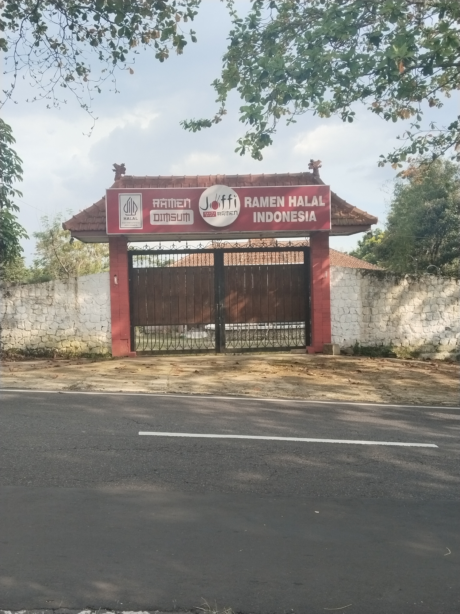The image presents the entrance to a business, identified as a "Ramen Halal Indonesia" establishment, located by an asphalt road in Karangpandan, Indonesia. The main subject is a large gate structure, featuring a red-tiled roof with traditional Indonesian architectural details on its peak. A prominent red rectangular sign is affixed to the front of this gate, above a set of closed, dark brown double gates.
The sign displays several pieces of text and logos. On the left, a white "HALAL" logo is visible, accompanied by smaller text beneath it that reads "01311099270723 JOFFI RAMEN". To its right, "RAMEN DIMSUM" is written in white capital letters. Centrally on the sign, a circular white logo features the brand name "Joffi RAMEN", with Japanese characters above it. The far right section of the sign boldly declares "RAMEN HALAL INDONESIA" in white capital letters.
Flanking the gate are low, rough-hewn stone walls painted white, suggesting a boundary or enclosure. Beyond these walls, various trees and foliage are visible. In the foreground, an asphalt road stretches across the width of the image, featuring a white line marking a lane boundary. The ground directly in front of the gate and walls is unpaved, covered with dry leaves and soil. Overhead, branches with green leaves from large trees partially frame the sky, which appears bright and overcast. No people or vehicles are present, and no specific activity is taking place. The scene depicts a static view under daylight conditions, with diffused lighting and soft shadows.