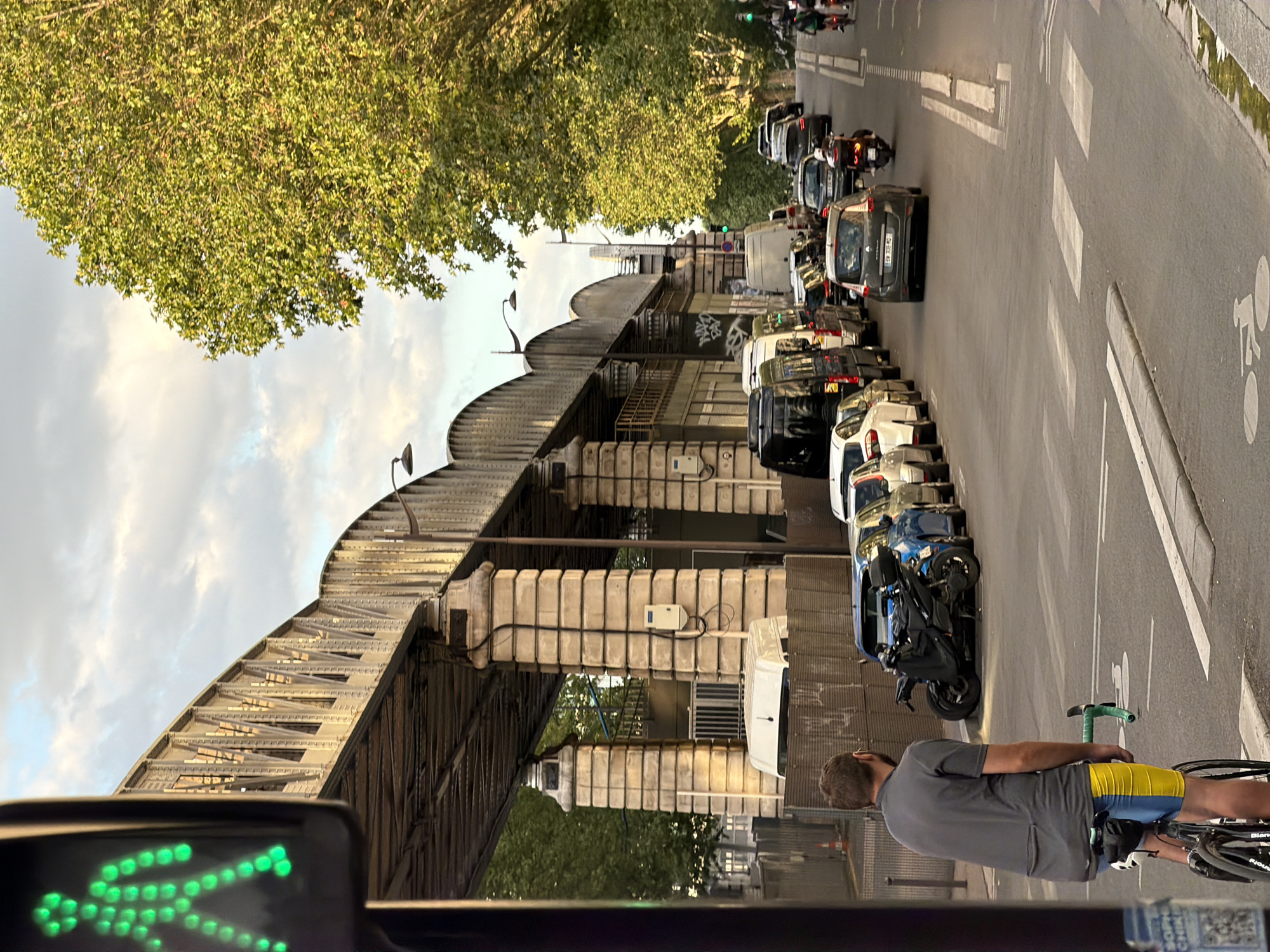 The image captures a street scene in Paris, France, beneath a distinctive railway bridge with arched girders. The bridge's robust stone pillars support the structure. A cyclist, a male wearing a grey t-shirt and blue and yellow shorts, is positioned in the foreground on the left, facing away from the camera. He is astride a bicycle with a green handlebar. A traffic light for pedestrians, illuminated with a green walking figure, is visible on the far left.
The street is lined with cars and motorcycles, indicating a period of traffic congestion. Among the vehicles are several vans, sedans, and at least one scooter with a rider. Trees with dense green foliage line the right side of the street, their branches extending towards the road. The sky above is partly cloudy. The overall scene depicts a typical urban environment with infrastructure, traffic, and pedestrian activity.