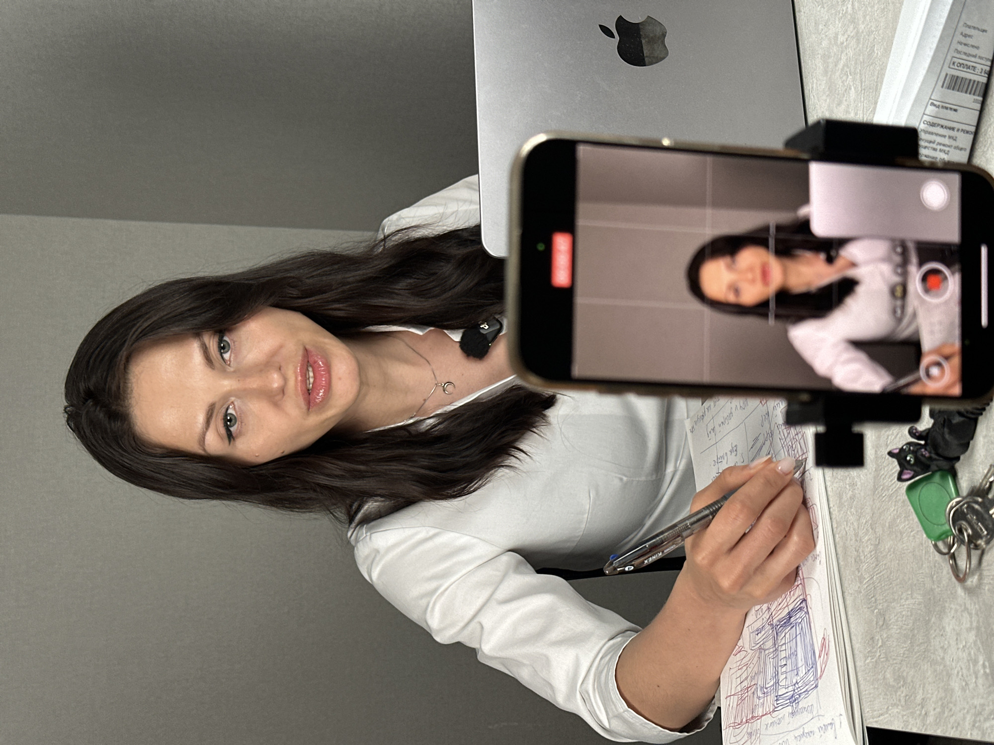 A medium shot captures a woman sitting at a desk, engaged in writing on a notebook. She is wearing a white button-up shirt and has dark hair, with a small microphone clipped to her collar. In front of her, a smartphone is propped up on a stand, displaying a recording of herself. To her right, a silver laptop with an Apple logo is visible, along with what appears to be a box of some sort. On the desk, near the bottom of the frame, are a set of keys attached to a green tag and a black cat-shaped keychain. The background is a plain gray wall. The woman is actively writing, holding a pen in her right hand and resting her left hand on the notebook.