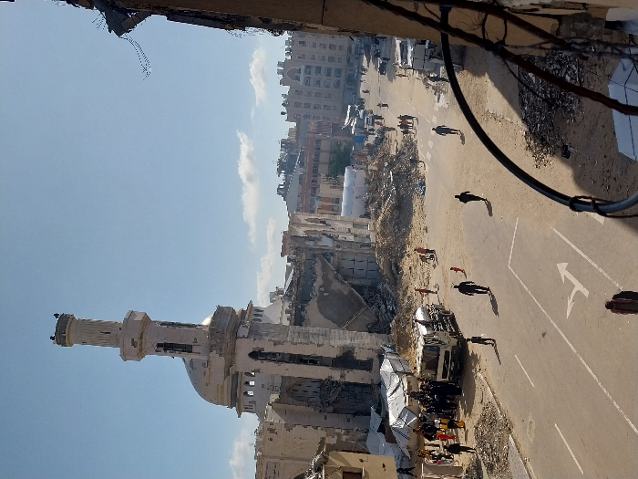 The image displays a wide view of a heavily damaged urban street in Khan Yunis, Palestinian Territory, under a clear blue sky with sparse clouds. The most prominent structure is the Great Mosque of Khan Yunis (Al-Omari Mosque) on the left, exhibiting extensive destruction; its white minaret remains largely upright but is significantly damaged, and the main mosque building behind it is reduced to a partial skeleton of walls and collapsed sections of its dome. The street is a paved road, partially covered with debris, and features a white painted right-turn arrow. Piles of rubble, concrete, and twisted metal from destroyed buildings line both sides of the street. On the left side of the street, adjacent to the mosque ruins, a white tanker truck is surrounded by a gathering of approximately 20-30 individuals, some carrying yellow jerry cans and buckets, suggesting aid distribution. Several white temporary shelters are set up near this gathering. Numerous damaged multi-story buildings are visible in the background and along the street. Approximately ten individuals are dispersed across the street, walking towards the background or across, including a figure in red. The immediate foreground on the right includes blurred structural debris, possibly from a collapsed building.