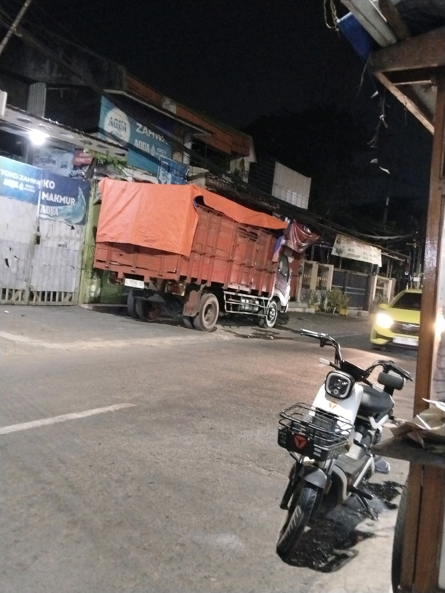 The image depicts a street scene at night in Surabaya, Indonesia. In the foreground, a white electric scooter is parked facing left, with its front basket visible. To the left of the scooter, a large red truck with an orange tarp covering its cargo is parked on the road. Behind the truck, several buildings with signs are illuminated. One sign displays "AQUA ZAMZAM". Further down the street, a yellow car with its headlights on is approaching. The environment appears to be a commercial or industrial area with streetlights casting illumination on the wet road.