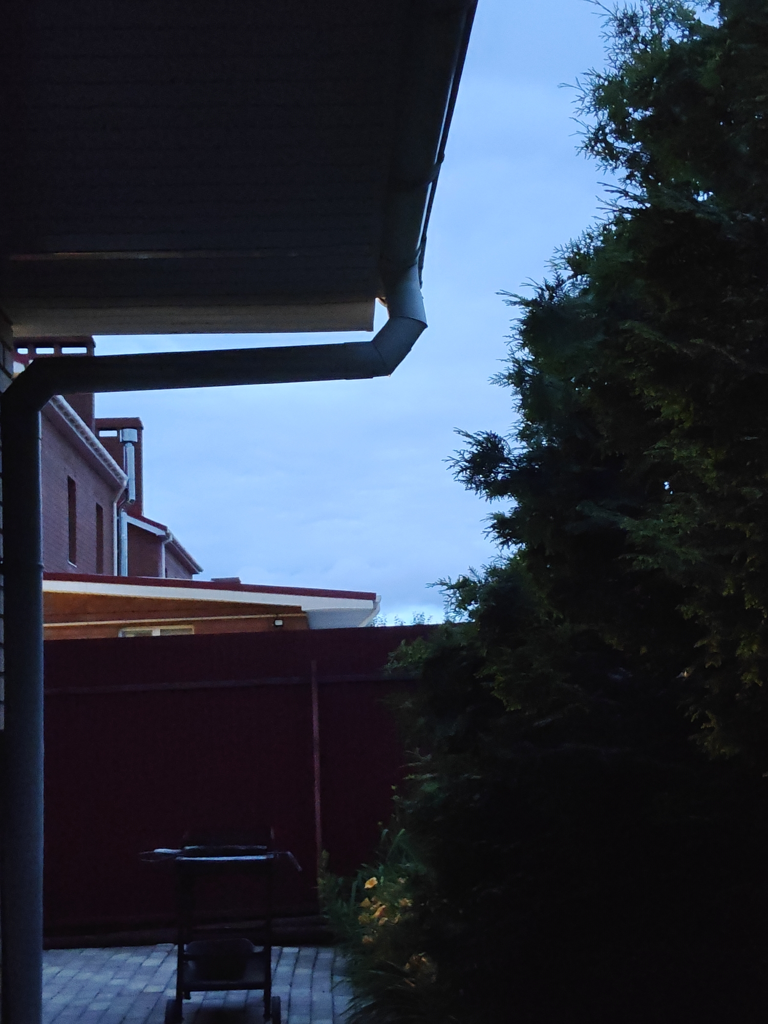 The image depicts an outdoor scene at dusk, likely in the city of Подстёпки, Russia. In the foreground, a dark evergreen tree dominates the right side of the frame, its foliage dense and casting a deep shadow. To the left, a portion of a dark-colored overhang or roof is visible, with a downspout extending downwards.
Beyond the overhang, a red brick house with white trim and red roofs is partially visible, suggesting a residential area. A dark red fence runs across the mid-ground of the image, separating the foreground from the structures behind it. In front of the fence, a black grill with a rack and wheels is placed on a paved patio made of interlocking bricks. Some yellow flowers are faintly visible to the right of the grill, near the base of the evergreen tree. The sky is a pale blue, indicating twilight or an overcast sky. No people are present in the image.