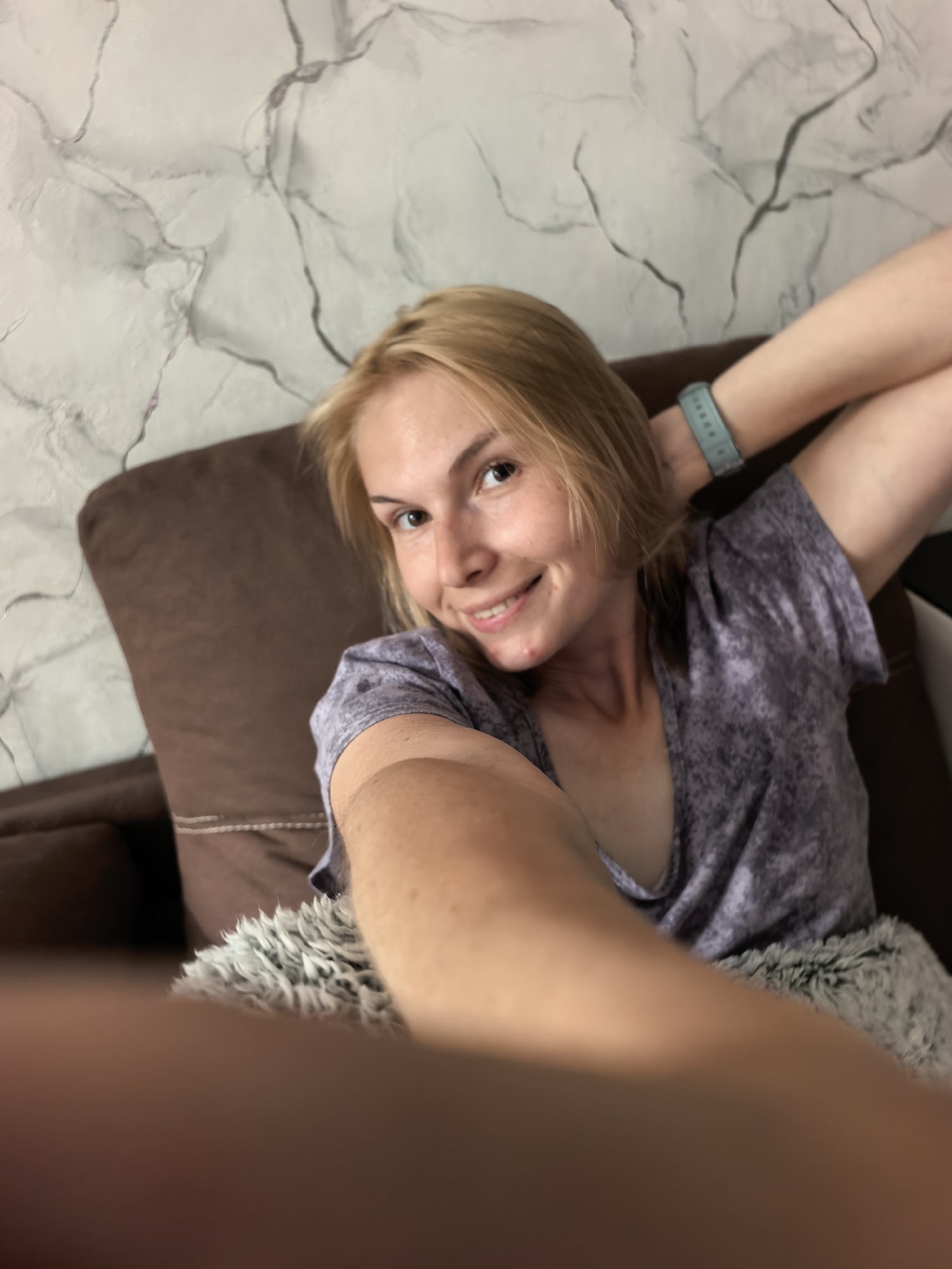 A young woman with blonde hair is taking a selfie indoors. She is reclined on a brown couch with a gray, fuzzy blanket. She is wearing a gray t-shirt and a blue fitness tracker on her wrist. The background shows a wall with a marble-like pattern. There is no evidence to confirm the location of the image is in Sterlitamak, Russia.