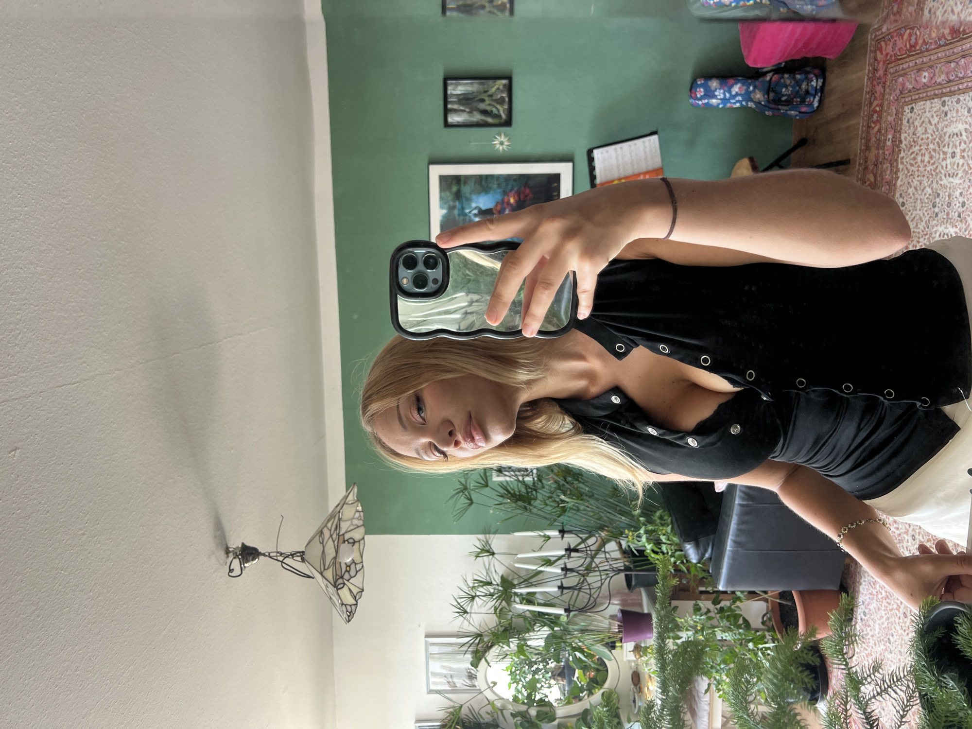 A woman with blonde hair and fair skin is taking a mirror selfie in a room. She is wearing a black sleeveless top with a V-neckline and white shorts. Her right hand is holding an iPhone with a transparent wavy case. The background shows a green wall with framed pictures and a large window. There are several plants, a chandelier, and furniture visible in the room. The lighting suggests it is daytime. The location is identified as Mühldorf a. Inn, Germany.