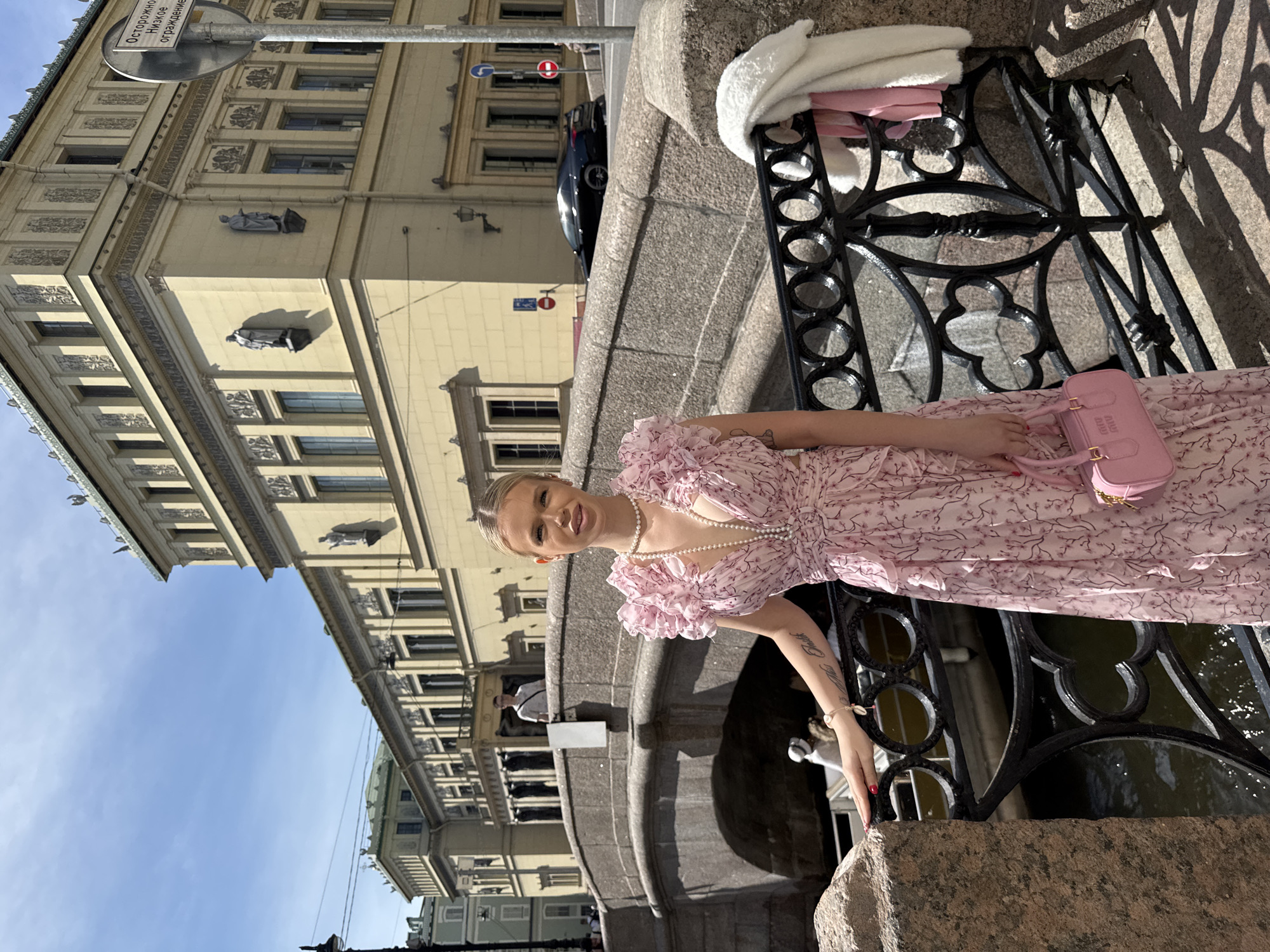 The media file shows a young woman posing by a canal in Saint Petersburg, Russia. The woman is wearing a pink floral dress and a pearl necklace. She is holding a pink handbag. The background features a building with classical architecture and a blue sky. The sun is shining, casting shadows on the scene. The overall mood is cheerful and relaxed, suggesting a pleasant day out.
The woman has blonde hair and appears to be in her early twenties. She is looking at the camera with a smile, her tongue slightly sticking out. Her left arm is resting on the canal's railing, and her right hand is holding the handbag. The dress is sleeveless with ruffled shoulders and a fitted bodice.
The building in the background is ornate, with windows and decorative elements typical of classical architecture. The canal water is visible below the railing, reflecting the sky and surrounding structures. The black metal railing is elaborate, with circular and curvilinear designs.
The time of day appears to be daytime, likely late morning or early afternoon, given the bright sunlight and shadows. The weather is clear and sunny, as indicated by the blue sky and the absence of clouds. The location is identifiable as Saint Petersburg due to the architecture and the presence of canals, which are characteristic of the city.
There is some visible text on a sign in the upper left corner of the image, which appears to be in Russian. The text is partially obscured, but some legible words include "Осторожно" (Caution) and "низкое ограждение" (low railing). This text adds a practical detail about the environment.