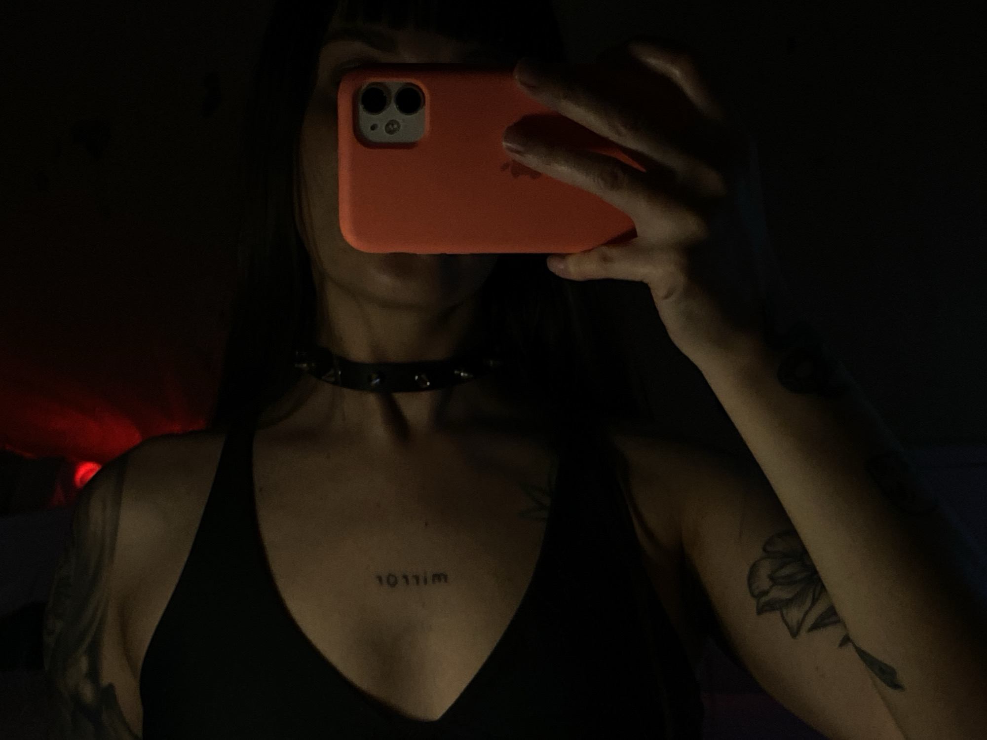 A dimly lit indoor selfie shows a person holding a salmon-colored iPhone in front of their face. The person has dark hair and is wearing a black top with a V-neck. A studded choker is around their neck. A tattoo of the numbers and letters "1011im" is on their chest. The left arm has a floral tattoo. The background is dark, with a red glow in the upper left corner. The location cannot be determined from the image.