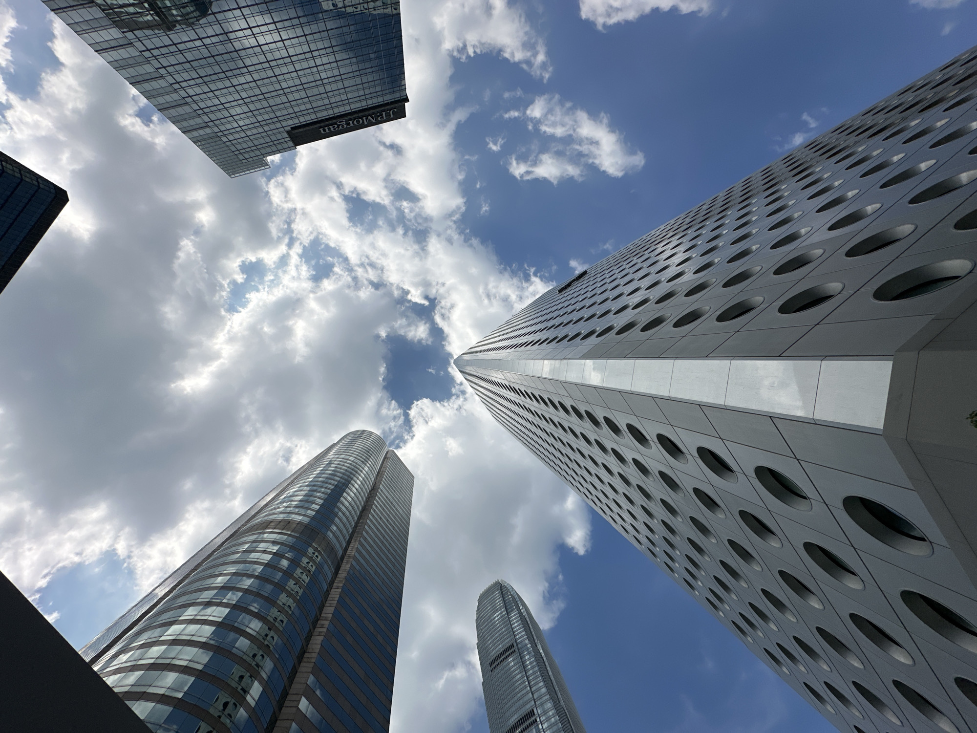 This is a low-angle shot of modern skyscrapers against a bright, cloudy sky in Hong Kong. In the foreground, a tall, white skyscraper with a facade of circular windows dominates the frame. To its left, a curved, glass-faced skyscraper reflects the sky and clouds. In the upper right corner, a dark skyscraper with a grid pattern of windows is visible, and the "J.P. Morgan" logo is discernible on its side. A fourth building, similar in style to the curved glass skyscraper, is partially visible on the far left. The image captures an architectural perspective of Hong Kong's financial district, emphasizing the geometric designs of its contemporary buildings and the expansive sky.