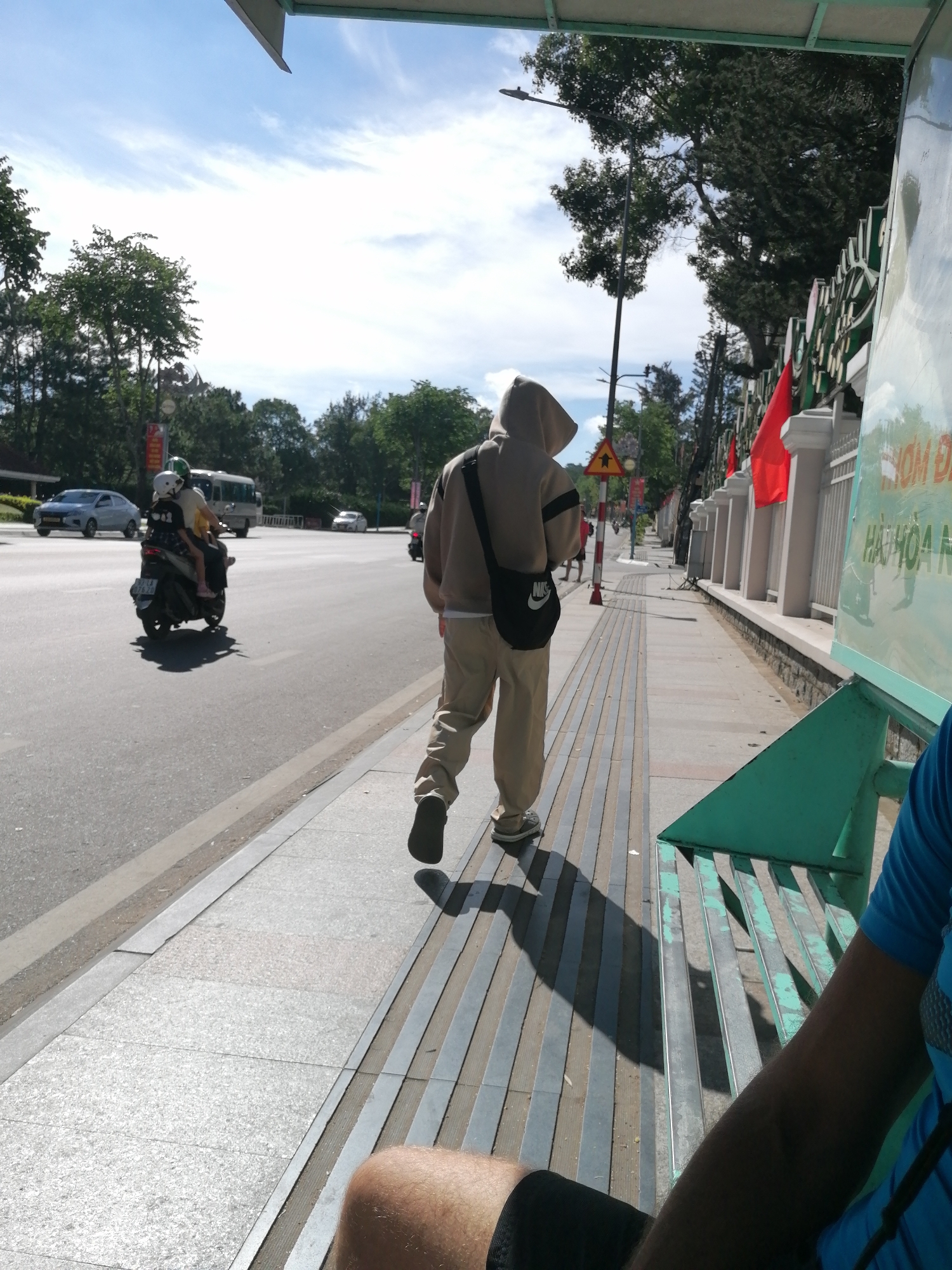 A person wearing a light brown hooded sweatshirt and beige pants walks along a sidewalk in Da Lat, Vietnam. They are carrying a black messenger bag with a Nike logo across their chest. To their left, a scooter with two passengers, a driver and a child, travels on the street. A white van and several cars are also visible on the road. The sidewalk is lined with metal railings and a green bench. In the background, trees and buildings can be seen. The sky is blue with some clouds.