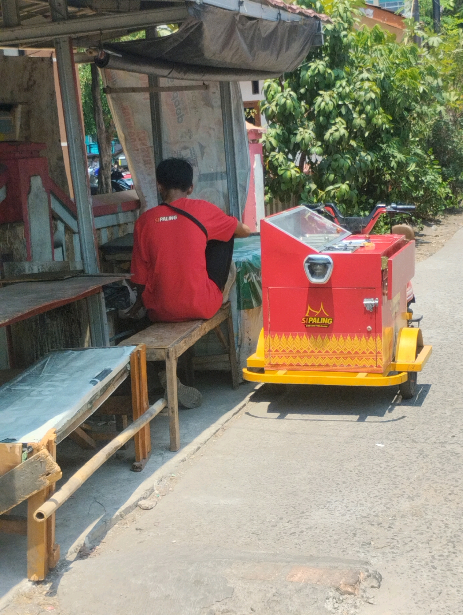 An outdoor scene shows a person wearing a red shirt with "SIPALING" printed on the back, sitting on a wooden bench. The person is positioned to the left of a bright red and yellow food cart with a glass display on top. The food cart is attached to a trailer with two wheels. In the background, there is a building with a dark roof and lush green foliage. The scene appears to be a street in Bekasi, Indonesia, given the distinctive red and yellow color scheme often associated with local vendors in the region, and the presence of tropical vegetation. The ground is paved with asphalt, and there is a wooden table structure in the foreground. The lighting suggests a bright, sunny day.