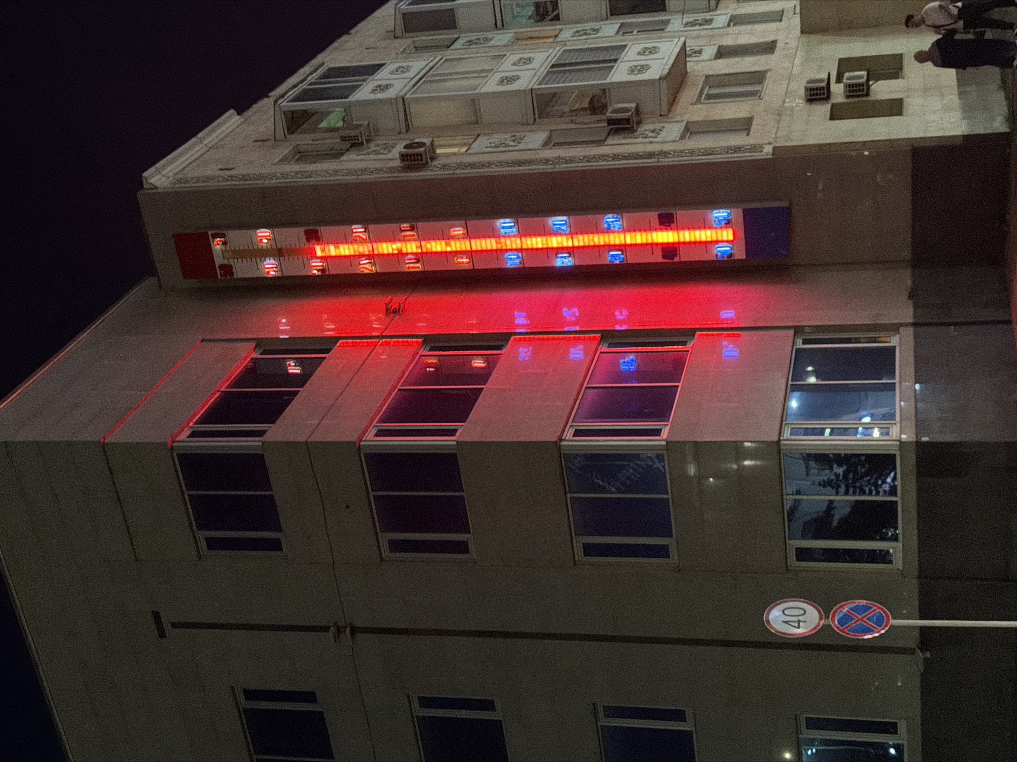 This image, taken at night in Pavlodar, Kazakhstan, depicts the exterior of a multi-story building, likely an apartment complex. A large, illuminated thermometer is affixed to the side of the building, displaying a red and yellow glow with blue indicators for temperature readings. The thermometer indicates a temperature of approximately 25 degrees Celsius. Two traffic signs are visible in the foreground: one shows a circular sign with the number "40" in a white circle with a red border, indicating a speed limit of 40 kilometers per hour, and below it, a circular sign with a red border and a blue cross, signifying no stopping or parking. The building features multiple windows, many with dark glass reflecting the surroundings, and some air conditioning units are mounted on the exterior walls. Two individuals, a woman in a dark headscarf and a man in a light-colored shirt, are standing near the base of the building, appearing to be conversing. The overall environment is urban, with the building and illuminated thermometer dominating the scene against a dark night sky.