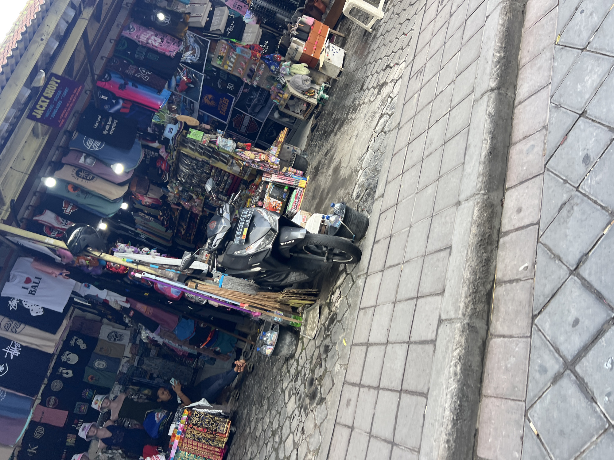 The image depicts a street scene in Kuta, Indonesia, likely a tourist area given the merchandise displayed. A busy street stall is the focal point, showcasing a variety of clothing, particularly t-shirts and hats, hanging from racks. Some of the t-shirts have text like "I Love Bali". A black scooter is parked in front of the stall, partially obscuring some of the merchandise. A person is seated on the ground to the left, looking at a mobile phone. The ground is paved with interlocking stones, and the stall is covered by a corrugated roof. The overall atmosphere suggests a vibrant marketplace.