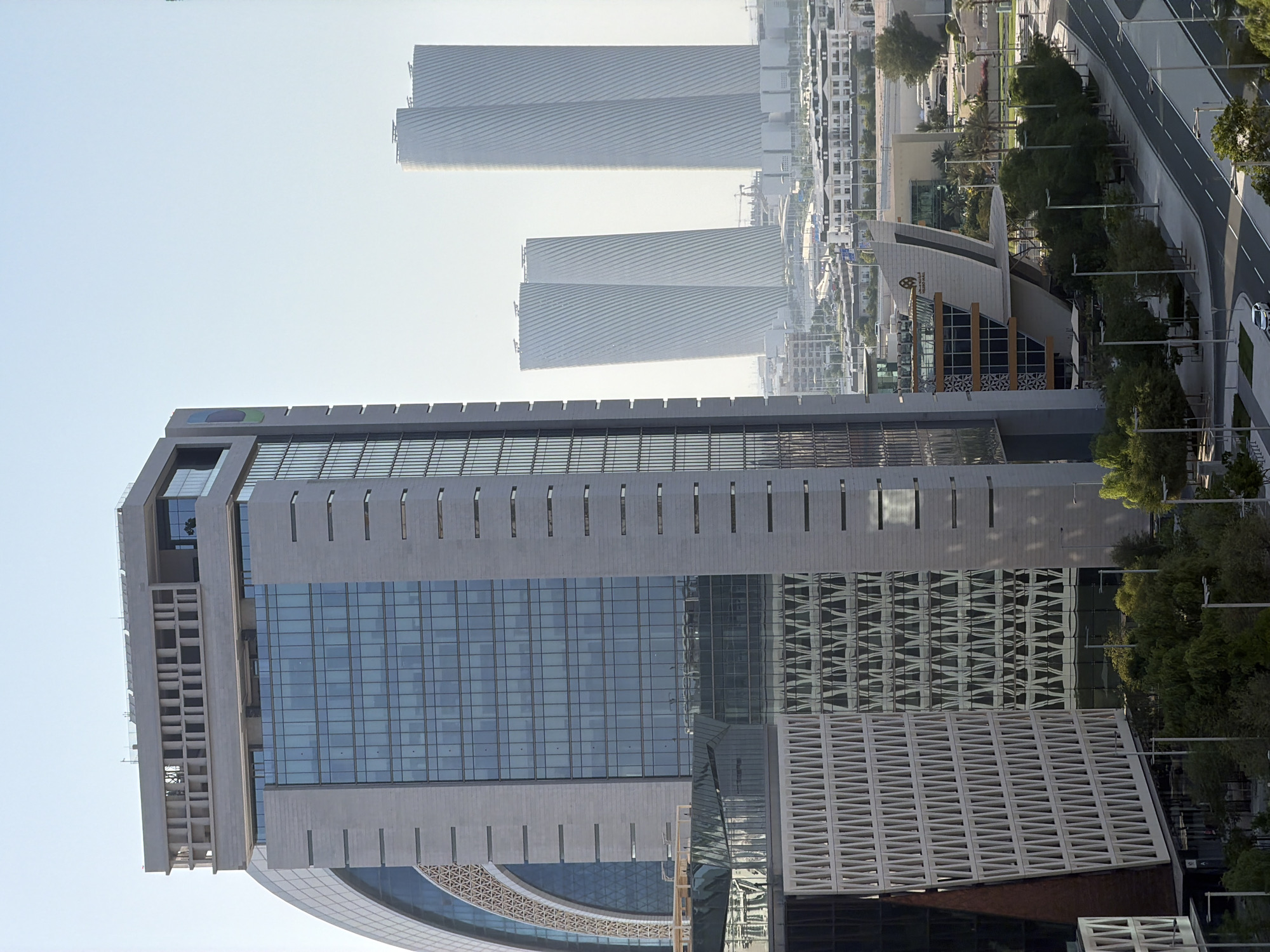 The image shows a view of a modern city with several prominent skyscrapers. In the foreground, a large, gray building with a facade of glass and concrete dominates the frame. The building features a large expanse of blue-tinted windows, with a patterned concrete structure on its lower section. Above and behind this building, other skyscrapers are visible, appearing as tall, slender structures with ribbed surfaces.
In the mid-ground, a distinctive building with a curved, angular design and a glass facade stands out. Trees and landscaping surround this building, and a road with a white dividing line curves alongside it, with a car driving on it. Further in the background, more buildings and city structures are visible, stretching into the distance under a clear, bright sky. The overall scene suggests an urban environment, possibly in a developing or modern city, with a focus on architectural design. The lighting indicates it is daytime, with no specific weather conditions noted. No people are clearly visible, and the activity seems to be general urban life. The image's orientation is rotated, making the sky appear at the sides.