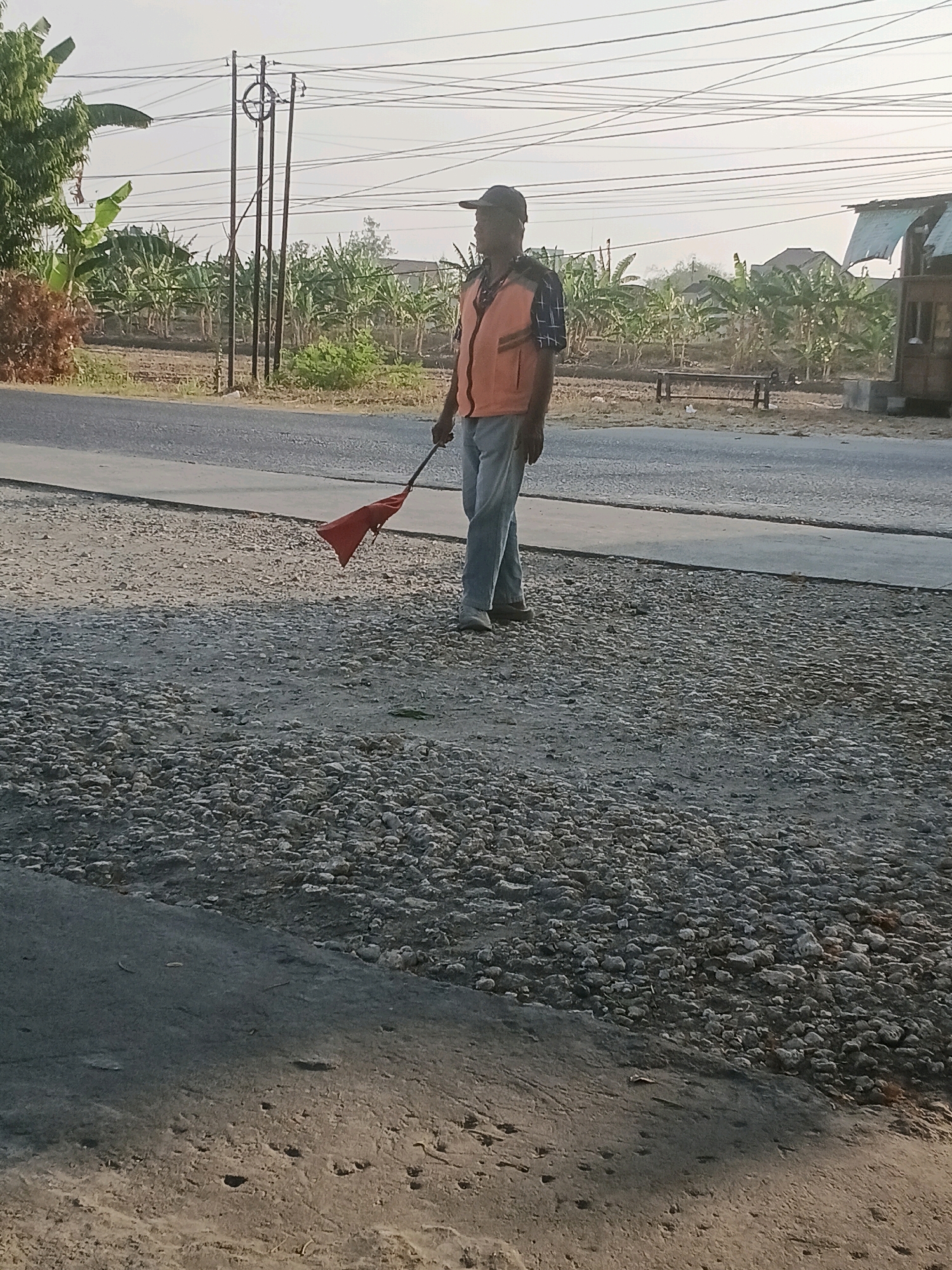 The image shows a man wearing a bright orange vest and a dark baseball cap standing on a gravel surface next to a paved road. He is holding a red flag attached to a stick, which is pointed downwards. The man is looking to his right, in profile. In the background, there are banana trees, utility poles with wires, and buildings. The sky is overcast. The scene appears to be in a rural or semi-rural area. Based on the context provided, this image is located in the city of Purwodadi, Indonesia.
