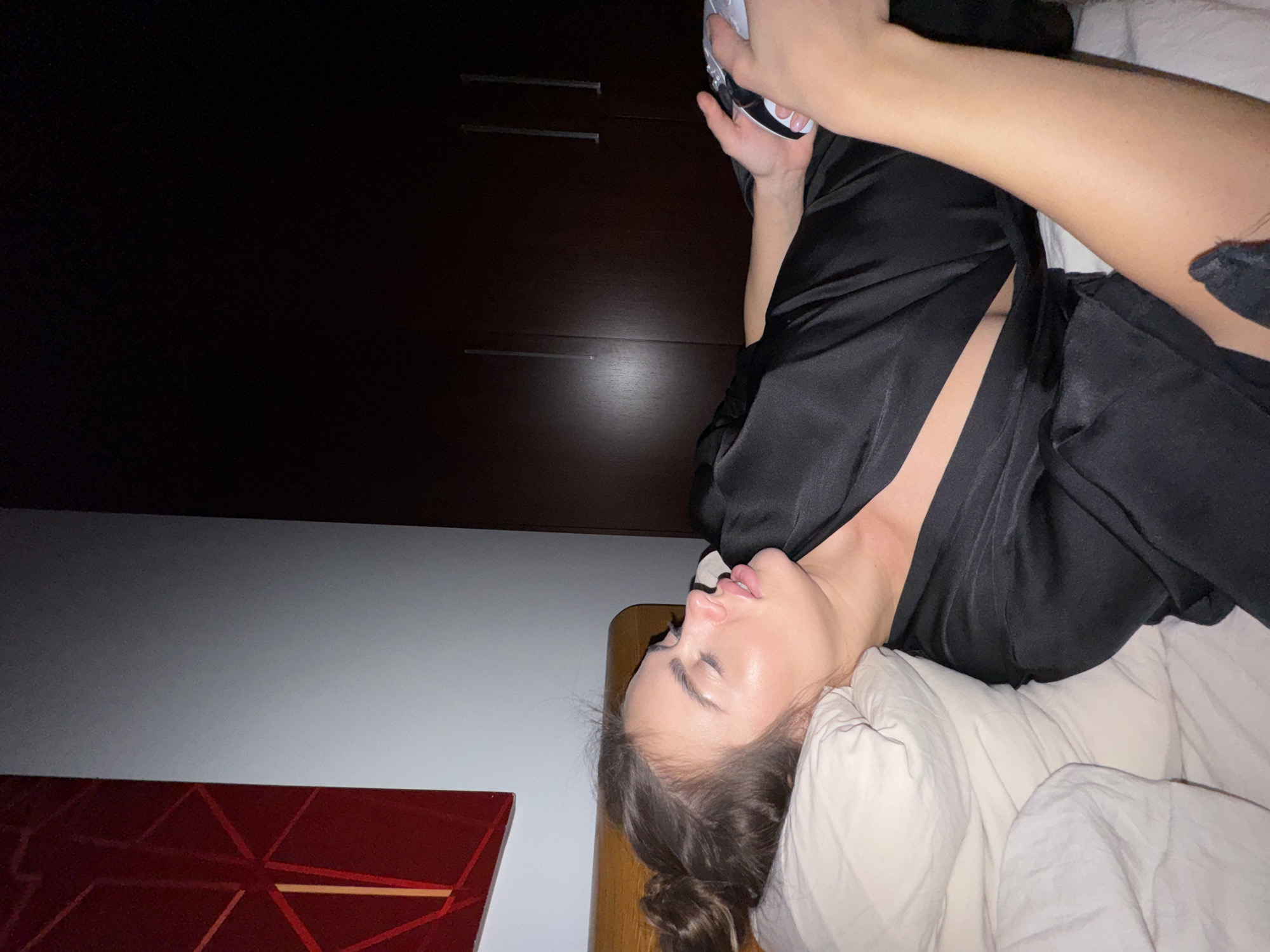 A woman lies in bed in an interior setting. She rests on white pillows and a bed with a wooden headboard. She is wearing a dark, satin-like garment that is partially open. She holds a white object in her hand above her. Behind her is a dark wooden cabinet with metal handles and a section of wall with a red geometric pattern on the lower left side. The location is unknown and cannot be determined to be in Warsaw, Poland based on the information in the image.