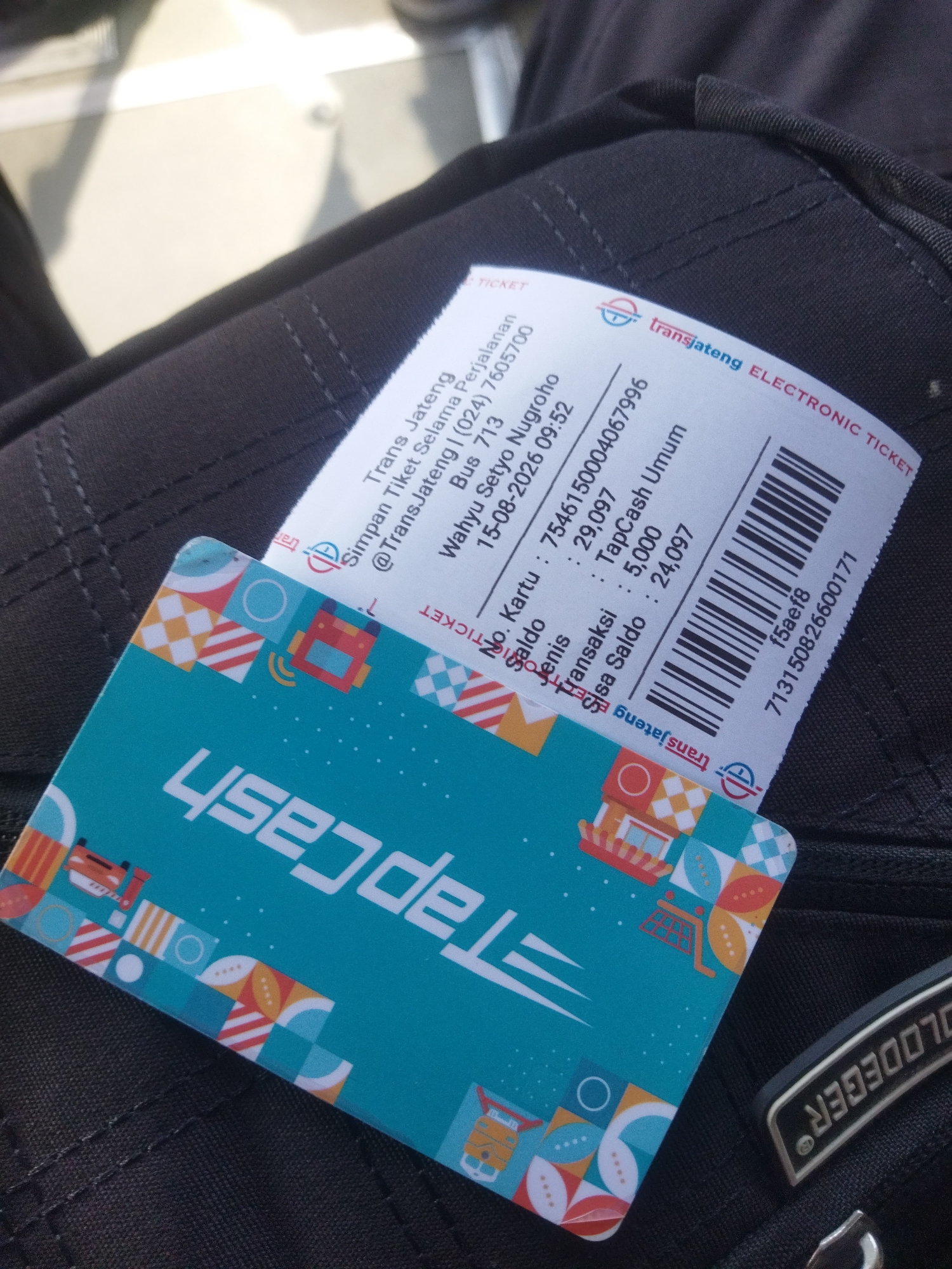 The image shows a close-up view of a Trans Jateng electronic ticket and a TapCash card on a dark, textured surface, likely a bag. The ticket is white with blue and red text and a barcode. It displays information such as "Trans Jateng," "Simpan Tiket Selama Perjalanan," "@TransJateng | (024) 7605700," "Bus 713," "Wahyu Setyo Nugroho," "15-08-2026 09:52," "No. Kartu: 7546150004067996," "Saldo Jenis: 29,097," "Transaksi: TapCash Umum," "Sisa Saldo: 5,000," "24,097," and "713150826600171." The TapCash card is teal and features white text that reads "TapCash" and various geometric patterns and illustrations of buildings and a train. The background is dark and out of focus. The image is a photograph, likely taken with a mobile phone. No people are visible in the image. The location is not explicitly stated in the image, but Trans Jateng operates public transportation in Central Java, Indonesia. Given the context, it is possible this image was taken in or near Wonogiri, Indonesia, where Trans Jateng services may be available.