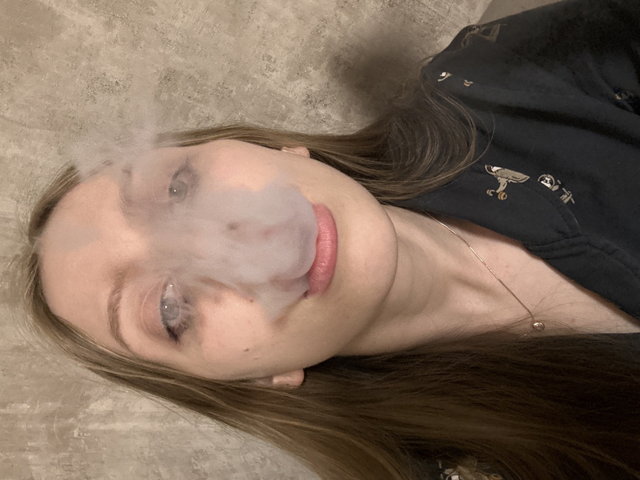 A close-up, chest-up indoor portrait of a light-skinned young adult female, located in Chaikovsky, Russia. She possesses long, light brown hair parted on the side and light-colored eyes. A cloud of white vapor or smoke emanates from her mouth, partially obscuring her nose and eyes. Her lips are slightly parted, revealing faint pink lipstick. She wears a dark-colored collared garment with a repeating pattern of light-colored abstract figures. A thin, silver-toned chain necklace with a small, teardrop-shaped pendant is visible around her neck. The background is a light brown, textured wall, suggesting an indoor environment.