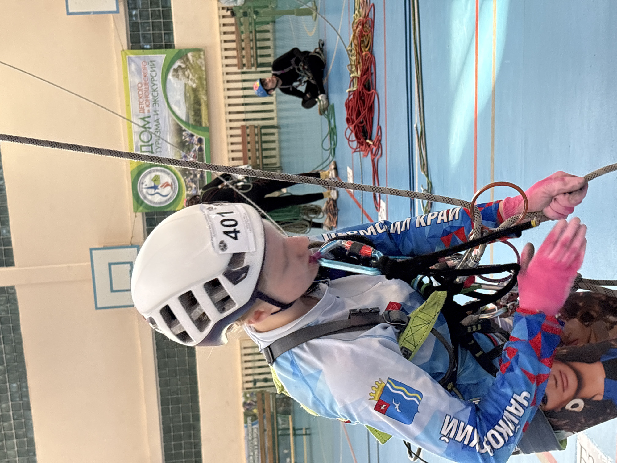A young female athlete, wearing a white helmet marked "401", a blue and white uniform with a regional flag patch, and pink gloves, is suspended on climbing ropes in what appears to be an indoor gymnasium. She is surrounded by climbing gear and ropes. In the background, another individual, also geared for climbing, is on the ground near more coiled ropes. A banner partially visible in the background advertises a "Home" for children's tourism and excursions. The uniform of the climber indicates affiliation with the Perm Krai region of Russia, suggesting the location is in or near that area. Given that the inscription on the sleeves of her uniform is written in Russian with the city of "ЧАЙКОВСКИЙ" visible, and the overall area is Perm Krai, it can be inferred that the location of the image is not in Berezniki, Russia, but likely within the Perm Krai region, specifically possibly related to the city of Chaykovsky. However, the city cannot be confirmed.