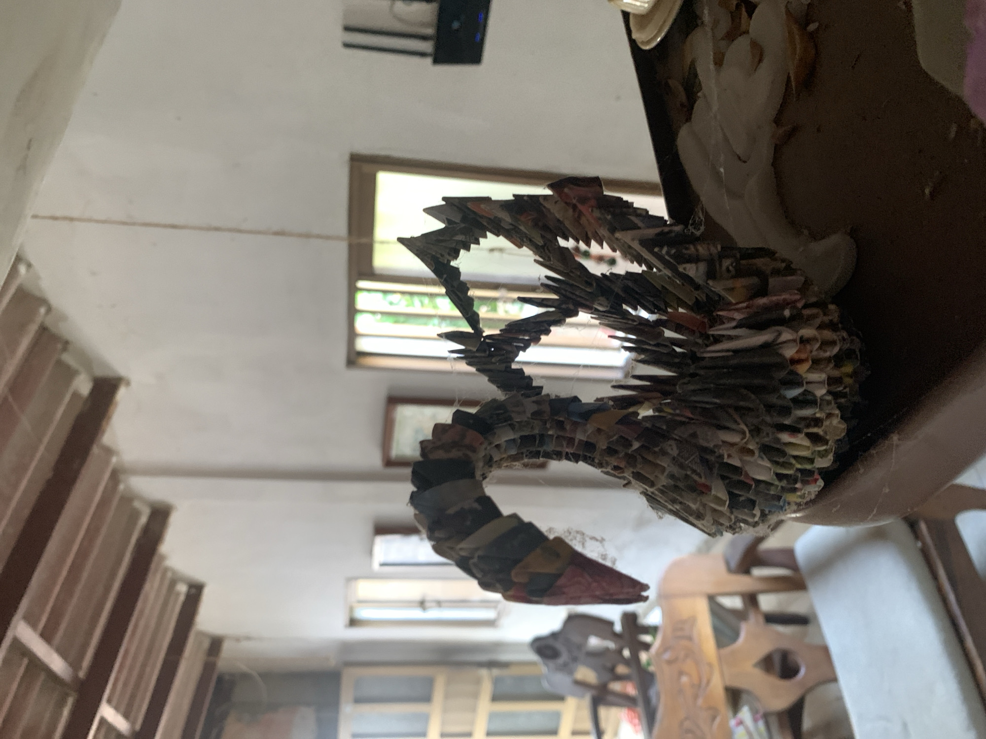 The image depicts a swan sculpture made of folded paper, sitting on a dusty table in what appears to be an interior space. The room has windows with natural light coming in, and there are chairs and other furniture visible in the background. The exact location in Santa Ana, Venezuela, cannot be determined from the image.