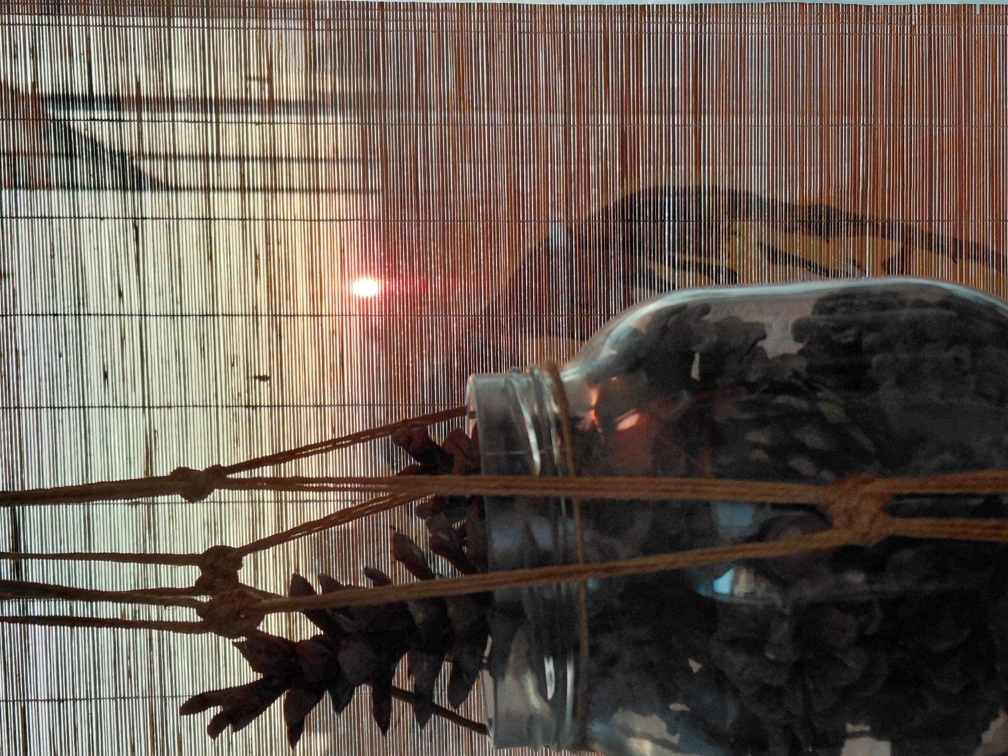 A close-up, eye-level shot shows a glass jar filled with pinecones, hanging by jute rope in front of a bamboo blind. The blind is partially open, revealing a sunset with a visible sun flare through the slats. The pinecones are dark and textured, with some tips extending above the rim of the jar. The jute rope is knotted in several places to create a hanger. The overall lighting is dim, with the sunset providing a warm glow. The scene is set indoors, with the window and blind suggesting a residential or commercial interior in Pleven, Bulgaria.