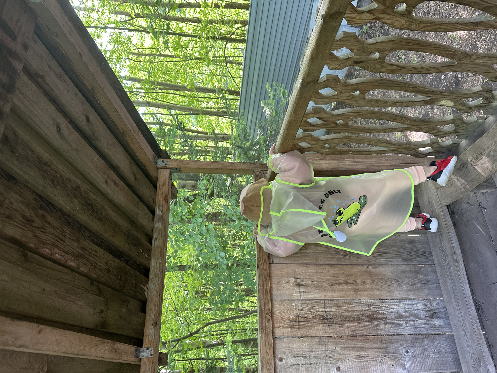 A child wearing a light pink outfit and a transparent raincoat with a cartoon frog on it is lying on their stomach on a wooden structure, possibly a lookout point or small building in a park. The structure has a carved wooden railing. The background shows green trees and a glimpse of a building. The location is Nizhny Novgorod, Russia.