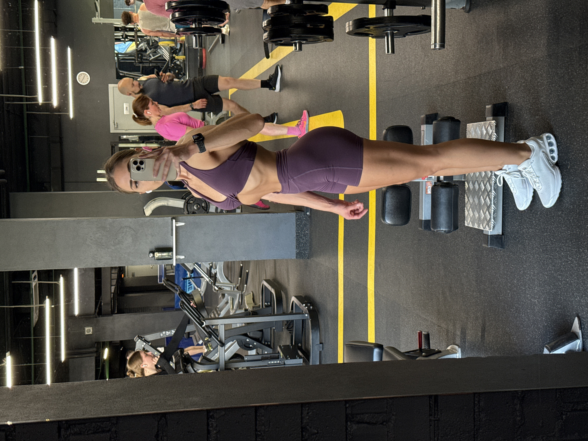 A young woman is taking a mirror selfie in a gym. She is wearing a purple sports bra and matching shorts, and white athletic shoes. Her hair is pulled back in a ponytail. In the background, other gym-goers are visible, along with exercise equipment. The gym appears to be well-lit and spacious, with dark walls and a dark floor marked with yellow lines. A clock is visible on the wall. The image is taken from the waist up, with the woman's body taking up most of the frame. The gym appears to be in Saint Petersburg, Russia.