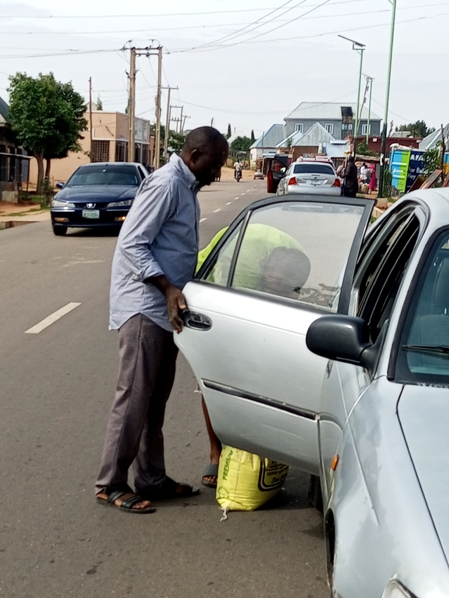 A man in a blue striped shirt and grey trousers stands on a paved road in Tempure, Nigeria. He is reaching into the open driver's side door of a silver sedan. A person wearing a lime green shirt is visible inside the car, bending down. A yellow bag with "PREMIUM" printed on it is on the ground by the car's rear passenger door. In the background, several buildings, trees, and utility poles are visible. Another dark blue sedan is parked on the road to the left of the silver car. The sky is overcast.