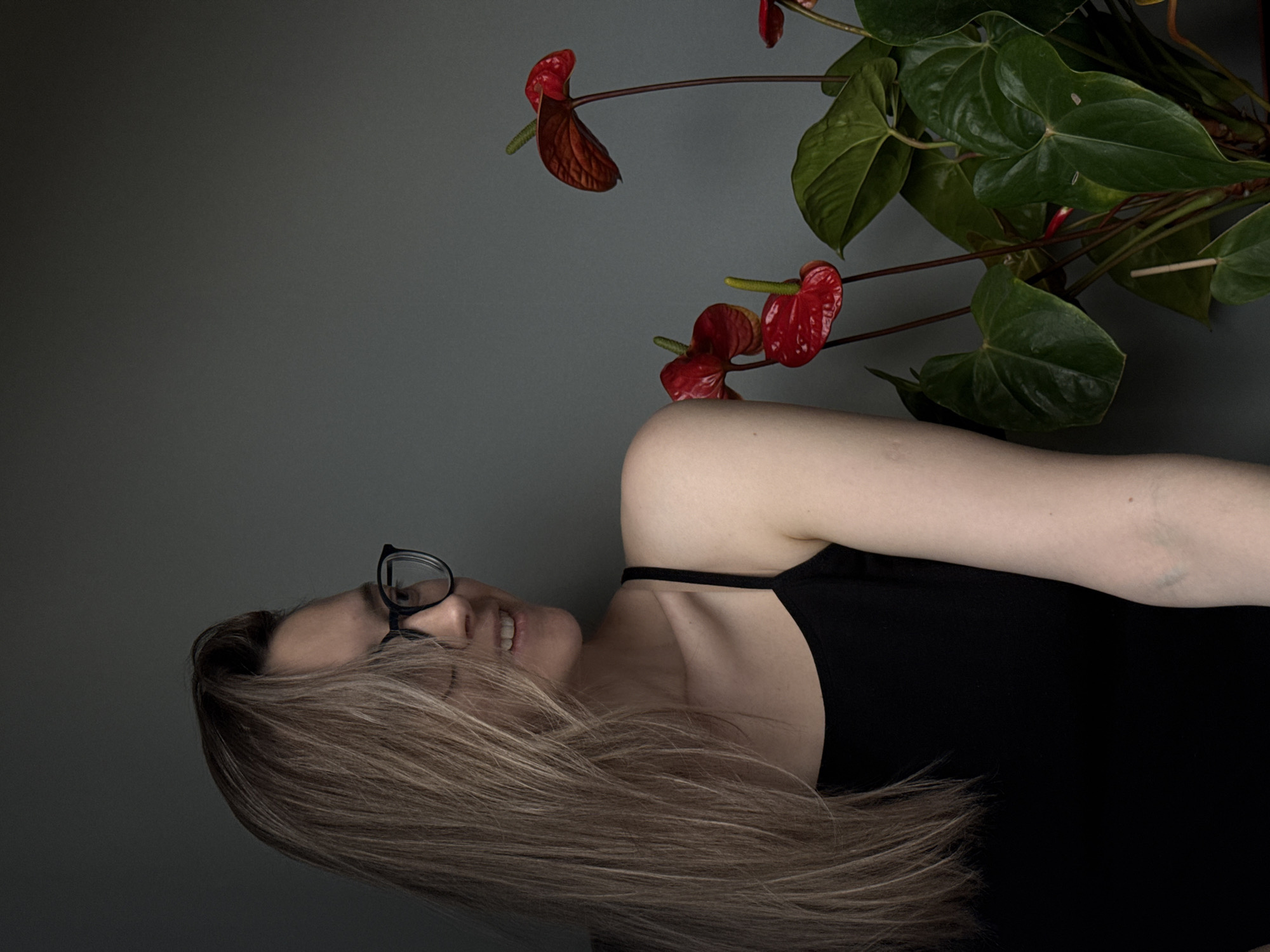 A portrait-style image, presented on its side, features a woman and an Anthurium plant against a plain, dark gray background.
The woman is positioned in the lower and central part of the frame (when upright). She has light skin, long, straight blonde or light brown hair that cascades downwards and to her right. She wears black-framed glasses and a black strappy top. Her face is in profile, looking upwards with a subtle smile or smirk, revealing a glimpse of her teeth. Her right arm is visible, bent at the elbow, suggesting a relaxed posture.
Above and slightly behind the woman, filling the upper right portion of the frame, is an Anthurium plant. It displays several vibrant red, heart-shaped spathes (often mistaken for flowers) with distinctive green spadices, accompanied by large, dark green, heart-shaped leaves. The plant's stems and leaves appear robust, contrasting with the muted background and the woman's dark clothing.
The lighting is soft and even, suggesting an indoor setting, possibly using artificial light or indirect natural light, highlighting the subjects without creating harsh shadows. There are no specific location cues visible, nor any discernible text. The overall mood is calm and contemplative, focusing on the interplay between the human subject and nature.