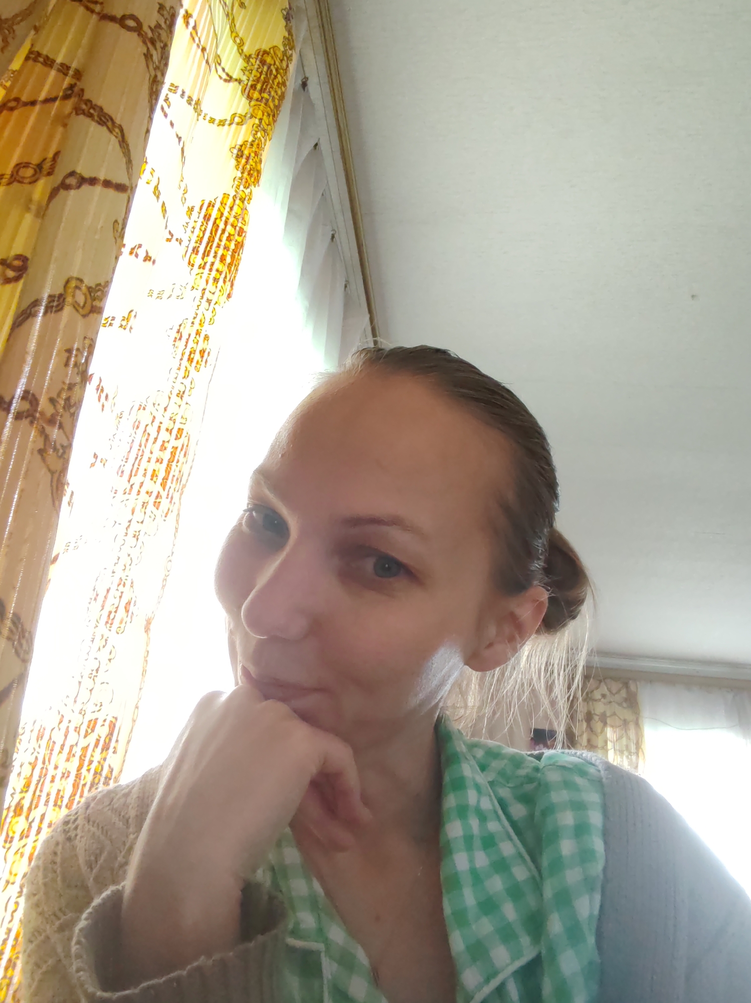 The image depicts a woman with fair skin and blue eyes, her hair pulled back in a bun. She is resting her chin on her hand. She wears a light-colored, possibly crocheted, sweater and a green and white checkered shirt or collar. The background features a window covered by yellow patterned curtains. The location is identified as Чухлэм, Russia.