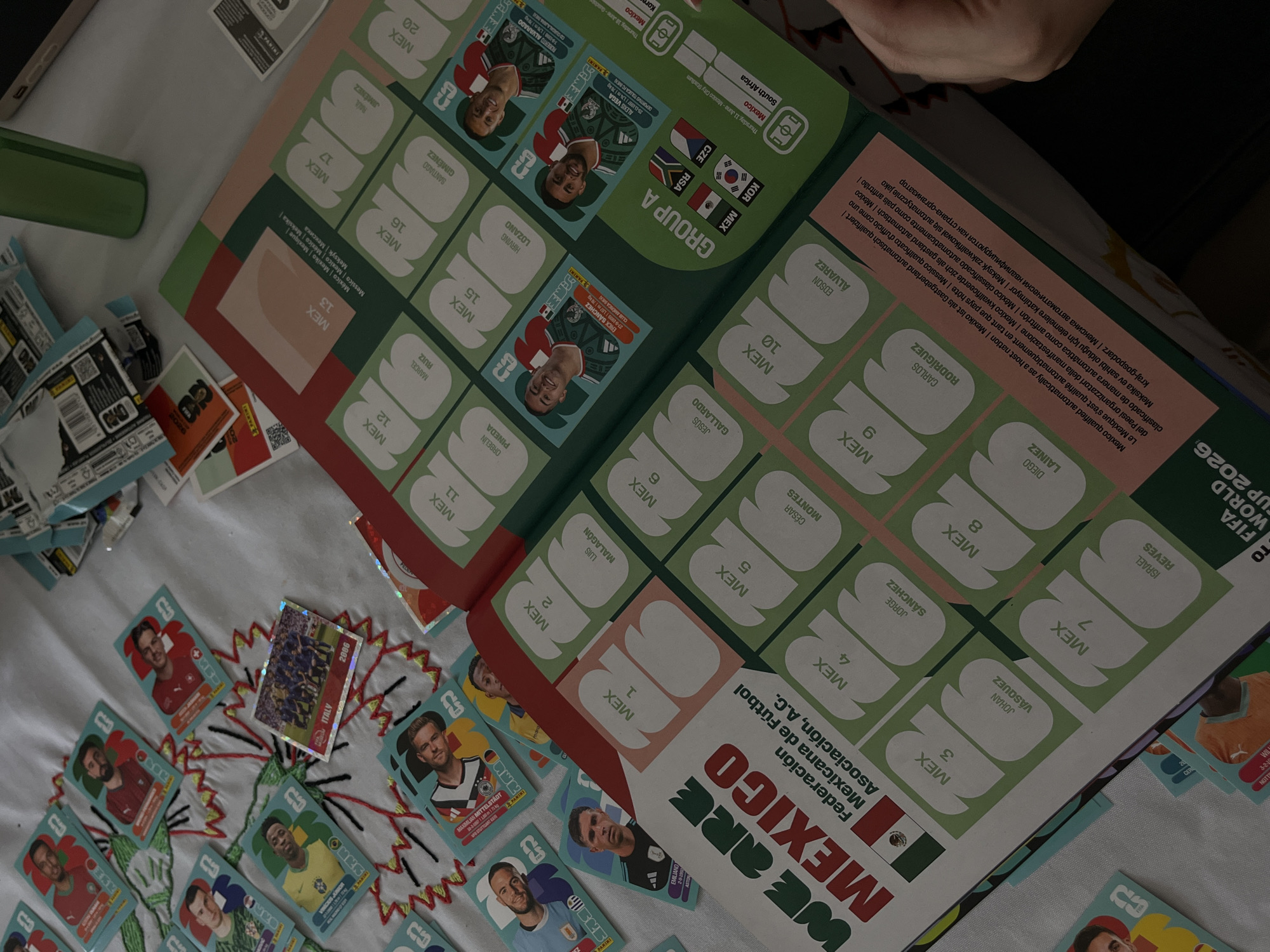The scene depicts a partially filled Panini Mexico 2022 FIFA World Cup sticker album. The open album shows the "We are Mexico" title page and the page for the Mexican national team. The album pages contain empty slots for stickers. Scattered around the album on a patterned surface are numerous loose Panini World Cup stickers featuring various soccer players from different countries. One sticker shows the Italian national team from 2006. The album also includes informational text about Mexico qualifying automatically as a host nation for the 2026 FIFA World Cup.