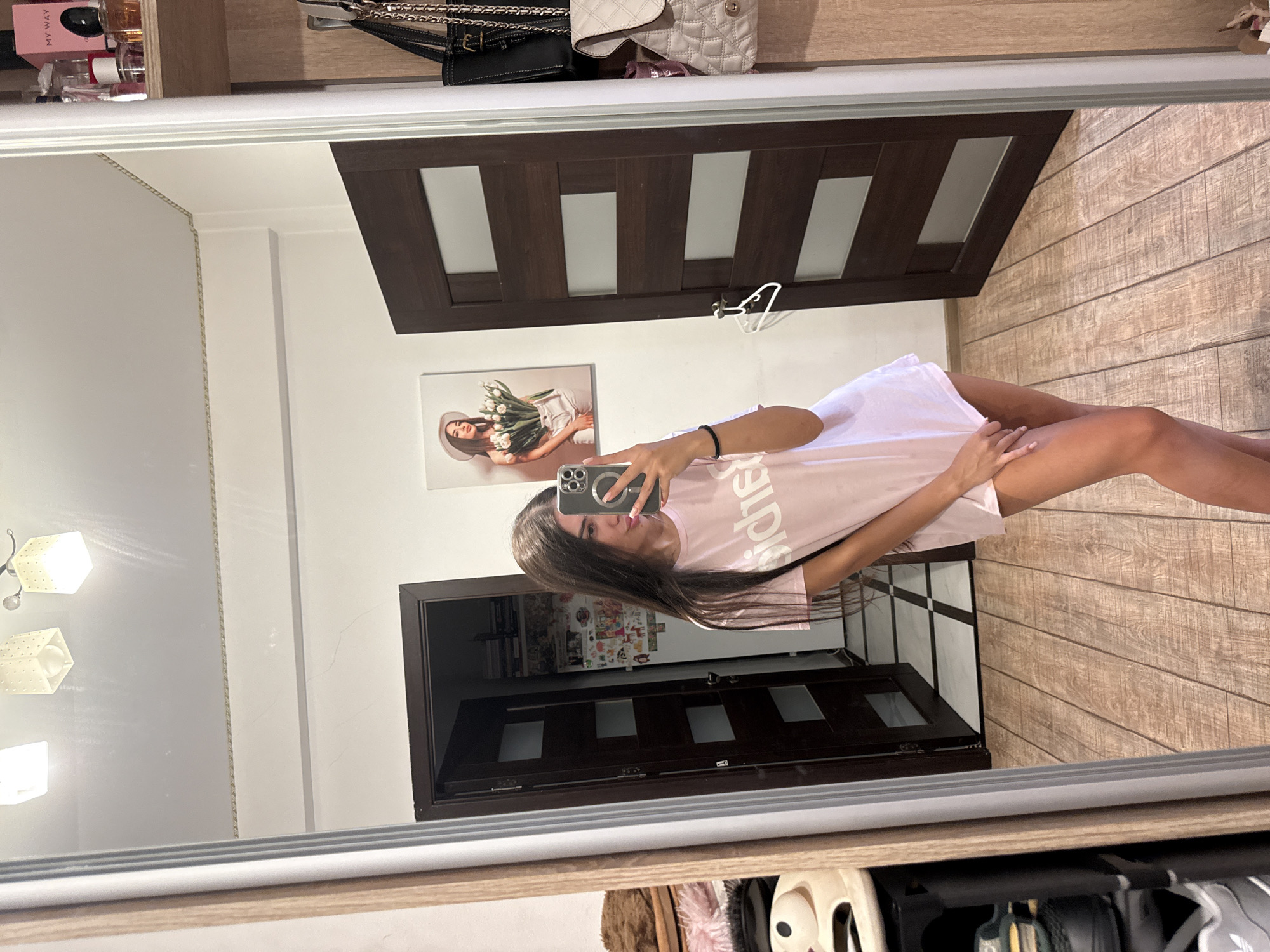A young woman with long, dark hair is taking a selfie in a mirror. She is wearing a pale pink oversized t-shirt and has her left hand on her hip. The background shows a room with wooden floors, a dark brown door with horizontal glass panels, and a shelf with perfume bottles. There is also a framed picture on the wall of a woman holding tulips. The reflection in the mirror also shows a closet on the left side with shelves and clothing. The scene appears to be an indoor setting, possibly a bedroom or dressing room. The location is identified as Chișinău, Moldova.
