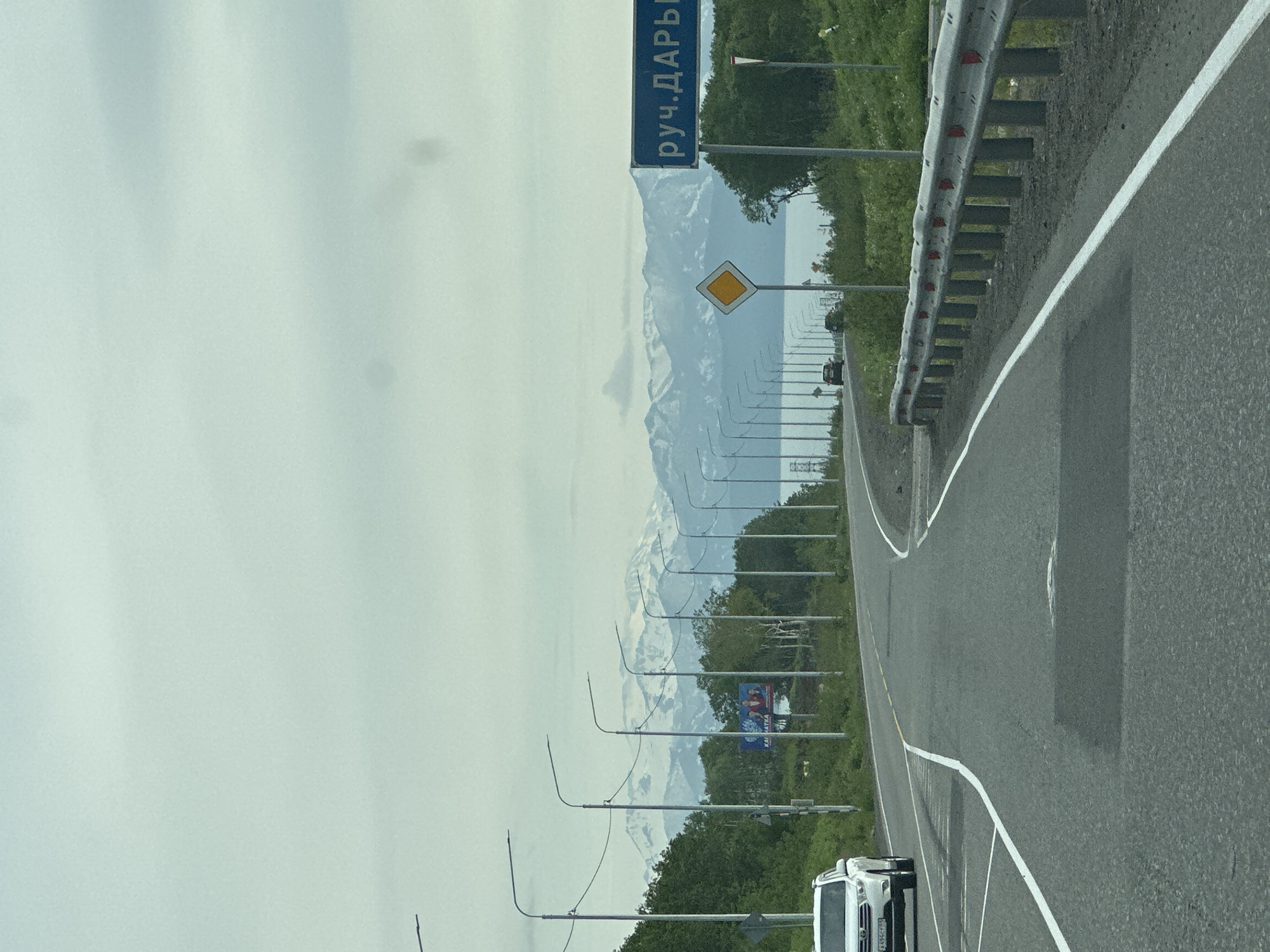 The image depicts a road scene in Petropavlovsk-Kamchatsky, Russia, with a focus on a highway leading towards a mountainous, snow-capped horizon under a cloudy sky. A white SUV is visible on the left side of the road. A blue road sign with white Cyrillic text, reading "руч.ДАРЫ", is prominently displayed on the right. Adjacent to the sign is a diamond-shaped yellow road sign. Numerous streetlights line the right side of the road, interspersed with green foliage. In the distance, a body of water can be seen beyond the trees. The road surface shows some wear and tear, with visible lane markings and a metal guardrail along the right edge.