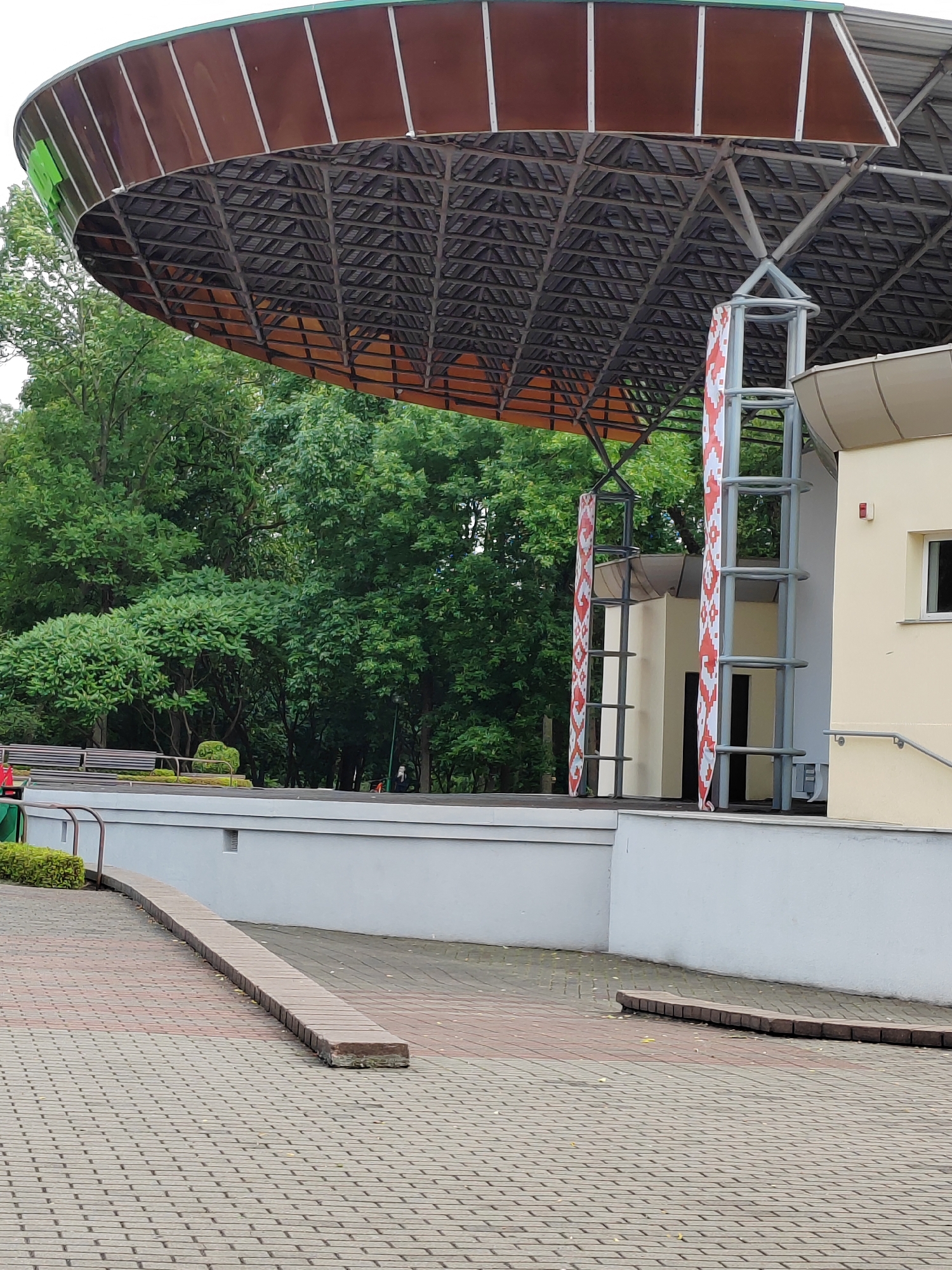 This is a medium-shot, eye-level photograph of an outdoor stage area. The stage has a large, curved, brown roof supported by a metal framework. The roof is partially covered with translucent panels, and the underside is a visible grid of metal beams. Red and white patterned banners are draped on either side of a metal support structure extending from the roof. To the right of the stage is a light yellow building with a small window and a railing leading up to an entrance. In the background, there are numerous green trees. The foreground consists of a paved area with a ramp-like feature made of interlocking bricks and a low, horizontal brick border. A bench is visible in the middle ground behind a low wall. The overall environment suggests a public outdoor space, likely a park or concert venue, in the city of Brest, Belarus. No people are clearly visible or identifiable in the image.