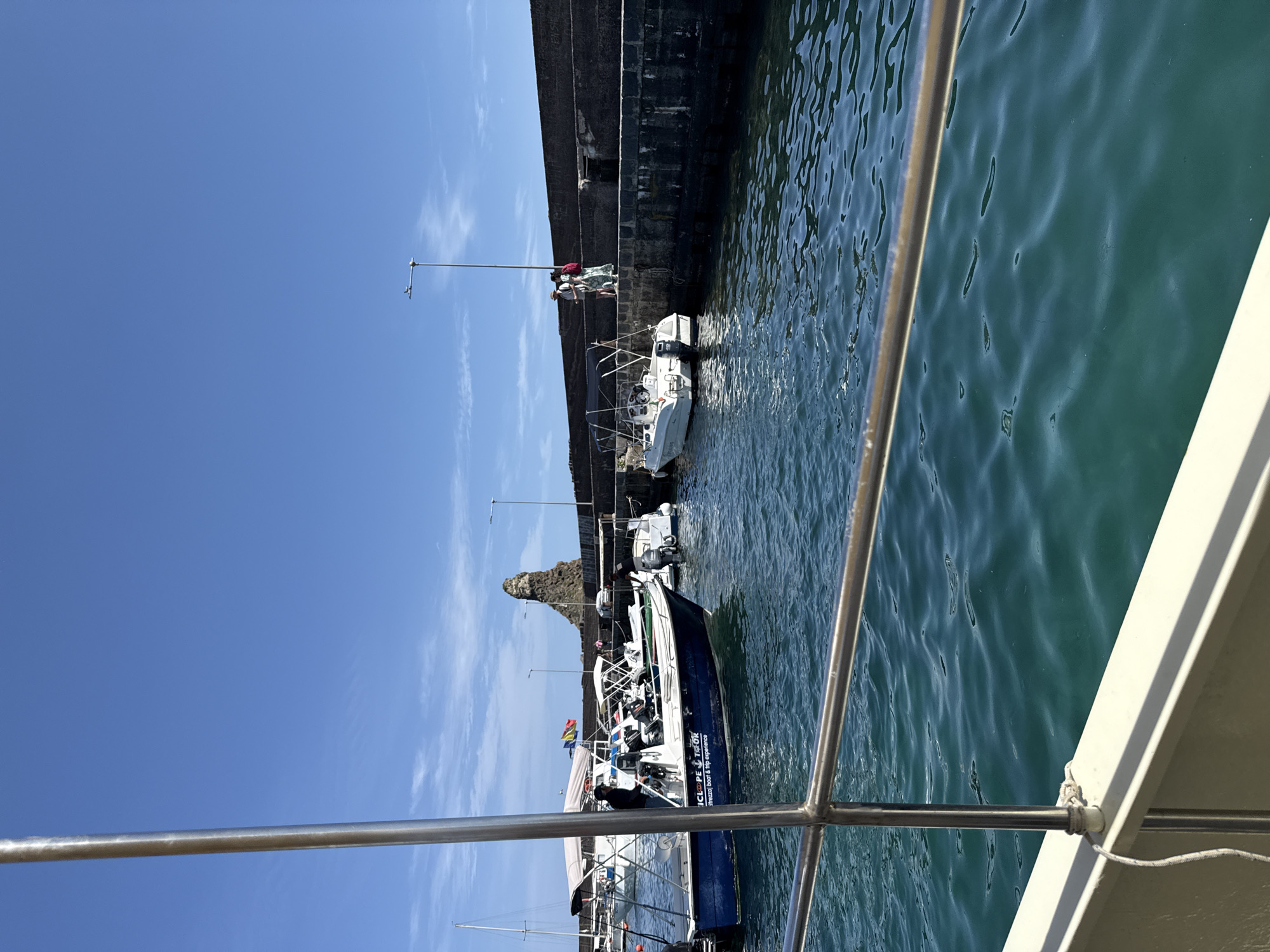 The image depicts a harbor on a clear day with a deep blue sky and light clouds. The water is a vibrant turquoise, marked by gentle ripples and reflections. A dark gray stone wall lines one side of the harbor, where several people are standing near boats docked alongside. The boats range in size from small sailboats to larger tourist vessels. One boat, closer to the viewer, has a visible banner with the text "CICLOPE Traozzl Boat & trip experiences". A rock formation rises near the harbor, adding to the scenic view. The perspective is from another boat, with parts of the boat's railing visible in the foreground, giving the viewer a sense of being on the water. The location is in the city of Unknown, Unknown.