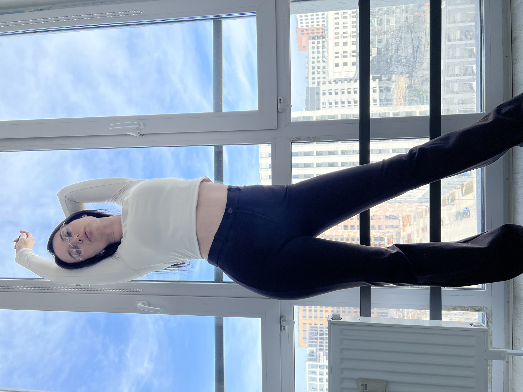 A woman stands in front of a multi-paned window, wearing a white long-sleeved top and dark pants. The background shows buildings, possibly in Tyumen, Russia, based on the urban landscape visible through the window. A radiator is visible near the bottom of the frame. The woman appears to be posing or stretching, with her arms raised and legs extended.