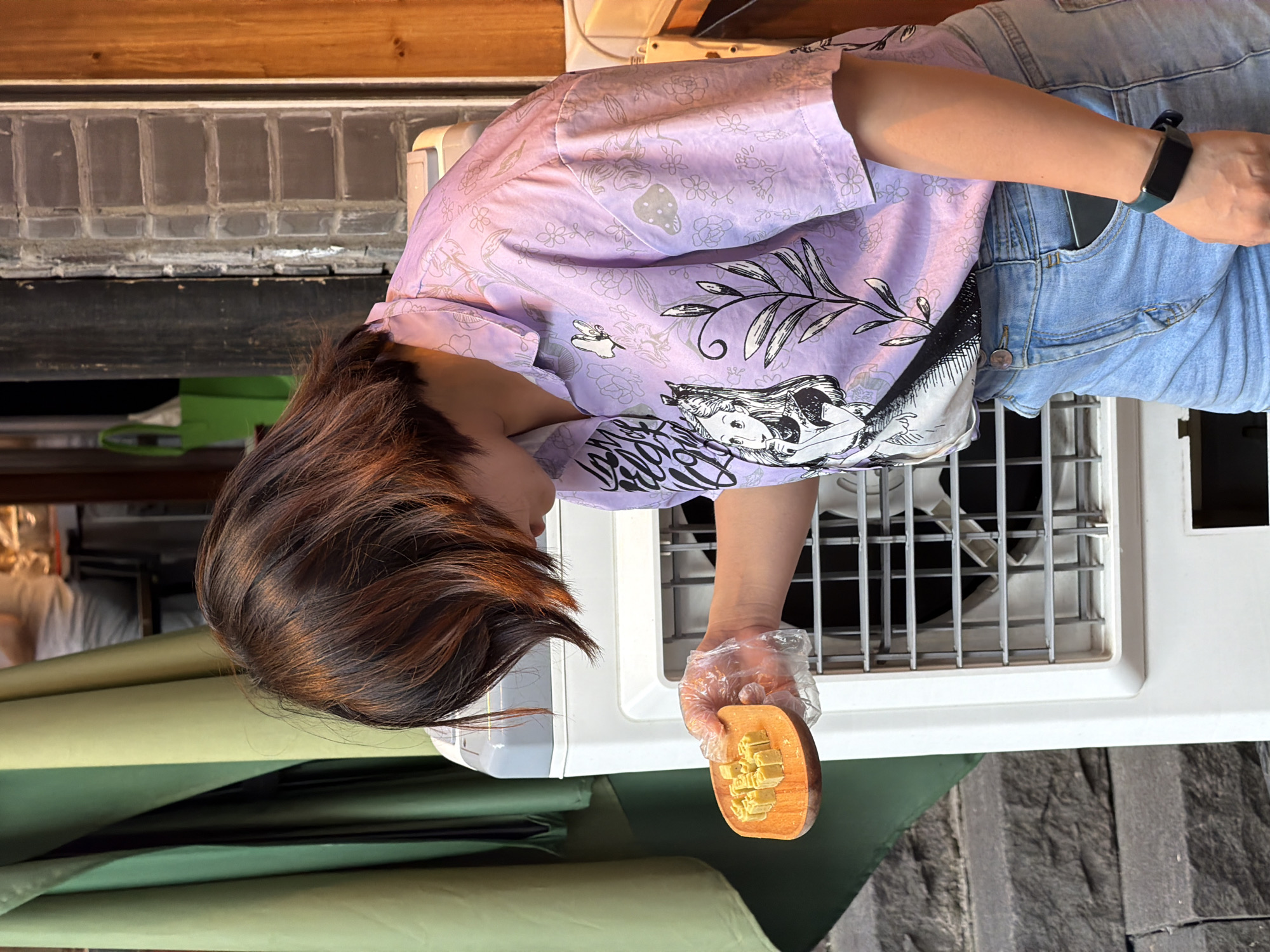 A young woman with short, reddish-brown hair stands in front of a white air cooler. She is wearing a light purple short-sleeved collared shirt with a pattern of Alice in Wonderland characters and floral motifs, and light blue denim shorts. The woman is holding a small, round wooden tray filled with several small, light yellow, cube-shaped candies. Her right hand, wearing a transparent plastic glove, is extended, holding the tray. The background shows a green curtain on the left, a brick wall and wooden paneling on the right, and stone steps leading down on the far left. The overall scene appears to be an outdoor street market or food stall, possibly in Yuzhong District, China, given the architectural style and the presence of a street vendor.