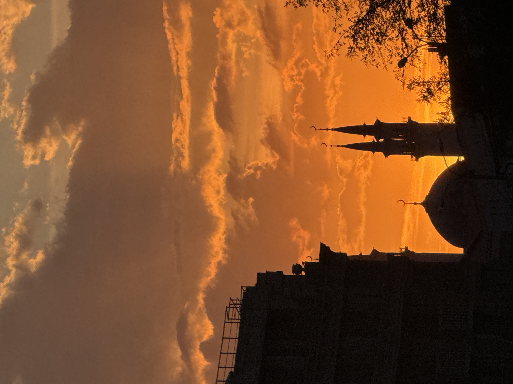 The image depicts the Qolşärif Mosque in Kazan, Russia, silhouetted against a vibrant orange sunset sky. The mosque's dome and two minarets with crescent moons are visible. In the foreground, a dark building with ladder is outlined. The sky is filled with textured clouds. A tree with thin branches and a street light are seen to the right. The image is oriented vertically.