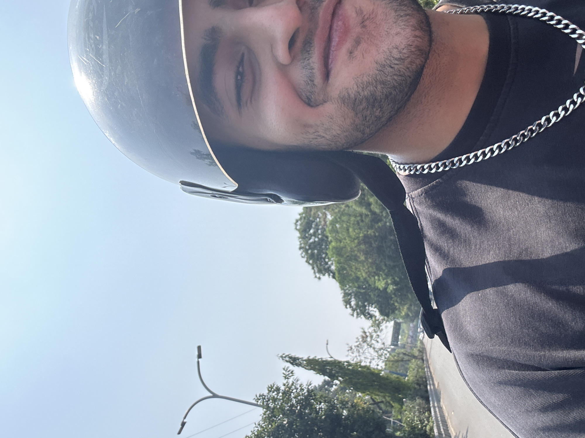 A close-up, tilted view depicts a man wearing a dark helmet and a silver chain, positioned against a backdrop of a clear sky and roadside trees. He has dark hair and a short beard. Trees and a light pole are visible in the background, suggesting an urban street. Based on visual cues and metadata, the location is likely Kolkata, India. The man appears to be either on a motorcycle or standing near the roadside.