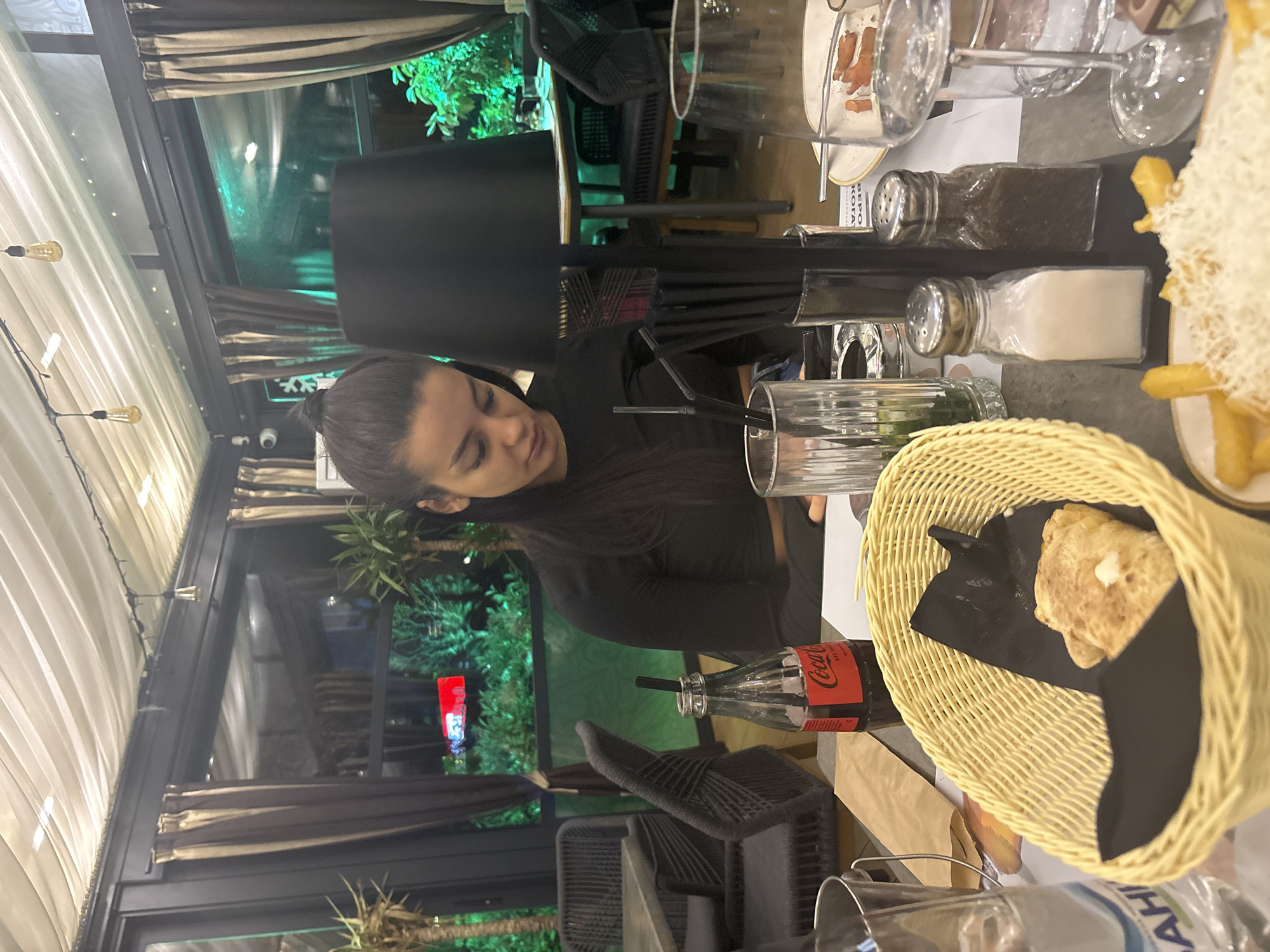 An image taken at a restaurant or cafe in Plovdiv, Bulgaria, depicts a dining scene during the evening. The photograph is oriented sideways, requiring a 90-degree clockwise rotation for correct viewing.
A woman with dark hair, tied back in a bun, is seated at a table, looking down. She wears a dark, long-sleeved top. The table is laden with dinnerware and food items, including a wicker basket holding bread on a black napkin, a plate of French fries topped with grated white cheese, salt and pepper shakers, and several glasses. A Coca-Cola bottle with a straw is also visible. A black lampshade stands on the table.
In the background, large windows or glass panels reveal an outdoor area with lush green foliage, illuminated with a greenish light, suggesting trees or plants outside the establishment. Striped curtains frame these windows. Overhead, bare Edison bulbs provide warm ambient lighting. A security camera is visible on the ceiling.
Visible text includes "Coca-Cola" on the bottle, partial text "BEPO KOJA" on a piece of paper behind the salt shakers, and "AHI" on a label at the bottom right. The setting suggests a casual yet comfortable dining environment.
