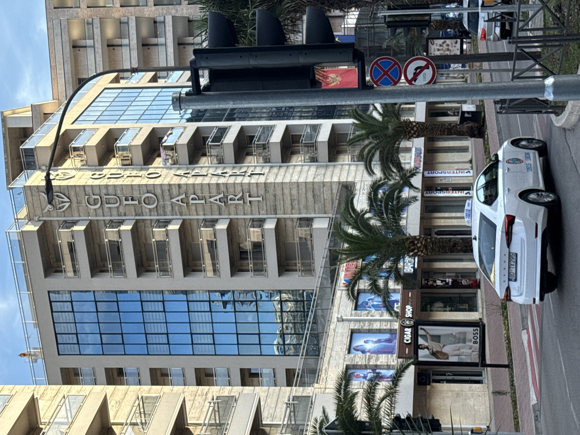 The image shows a street scene in Budva, Montenegro, featuring the "Gudzo Apart" hotel. The hotel is a multi-story building with a mix of glass and concrete facade. A street sign and traffic lights are visible in the foreground, partially obscuring the hotel's name. Palm trees line the street in front of the building, and various storefronts, including "INTERSPORT" and "CIGAR SHOP BOSS" are located on the ground floor. A white Skoda car with license plate "BD TX295" is parked on the street in front of the shops. The overall environment suggests a bustling commercial area in a coastal city.