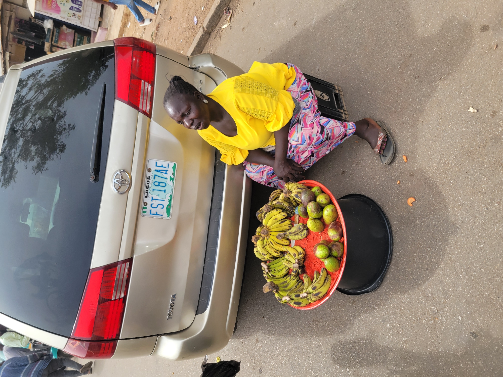 The image depicts an outdoor street scene in Jos, Nigeria, during daylight hours. The central subject is an adult woman with dark skin and braided hair, wearing a bright yellow short-sleeved top with embroidered details and a colorful patterned skirt or wrapper. She is crouching or kneeling on the ground next to the rear of a light gold Toyota minivan or SUV.
In front of her, a large red plastic basin, possibly sitting on a black plastic stool or crate, holds an assortment of fruits. These include several bunches of bananas, some yellow and ripe, others greener, and numerous green, oval-shaped fruits resembling avocados. This arrangement indicates she is a street vendor selling produce. Her facial expression appears focused or slightly strained as she attends to her wares. She wears simple flip-flop sandals on her feet.
The car's rear is prominently featured, displaying a Nigerian license plate with the text "LAGOS FST-187AE." The brand name "TOYOTA" is visible on the vehicle's rear hatch. The setting is a paved surface, likely a road or pavement, with a visible curb. In the background, other pedestrians and glimpses of buildings suggest an active, possibly commercial, urban environment. The lighting is bright and clear, indicative of a sunny day.