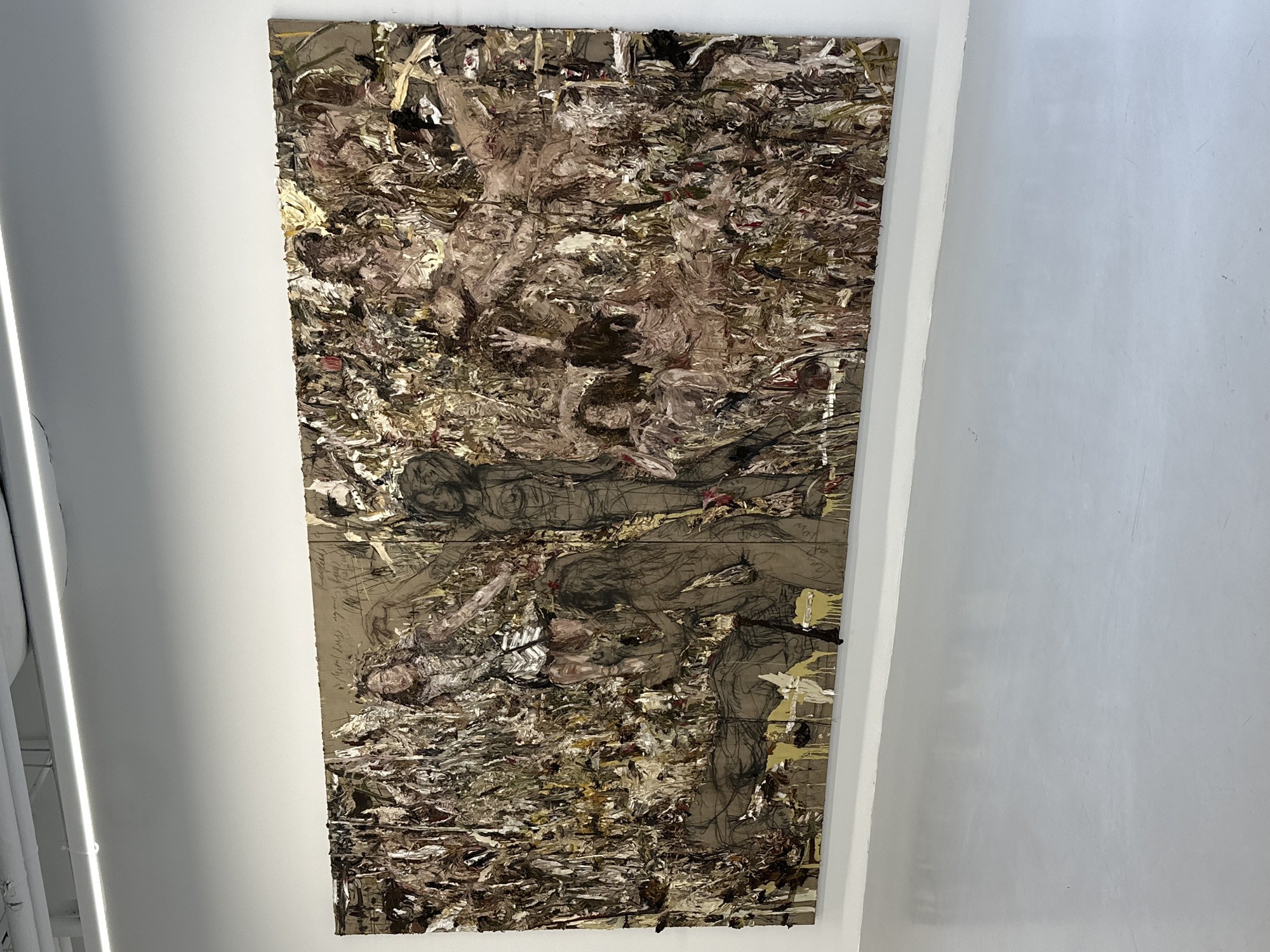 The image shows a large-scale artwork hanging on a white wall, likely in a gallery or museum setting in London, United Kingdom, given the architectural style of the building and general context. The artwork itself is a complex mixed-media piece, combining abstract expressionist painting techniques with figurative drawings. Heavy impasto textures in browns, creams, and golds dominate the upper and lower sections of the artwork. Interspersed within these abstract areas are discernible figures, rendered in a loose, sketch-like manner with dark lines and shading. The figures appear to be engaged in some kind of struggle or entanglement. The artwork is of significant size, spanning a considerable vertical length of the wall. To the left of the artwork is a portion of a modern lighting fixture attached to the ceiling. To the right of the artwork is an expanse of blank white wall.