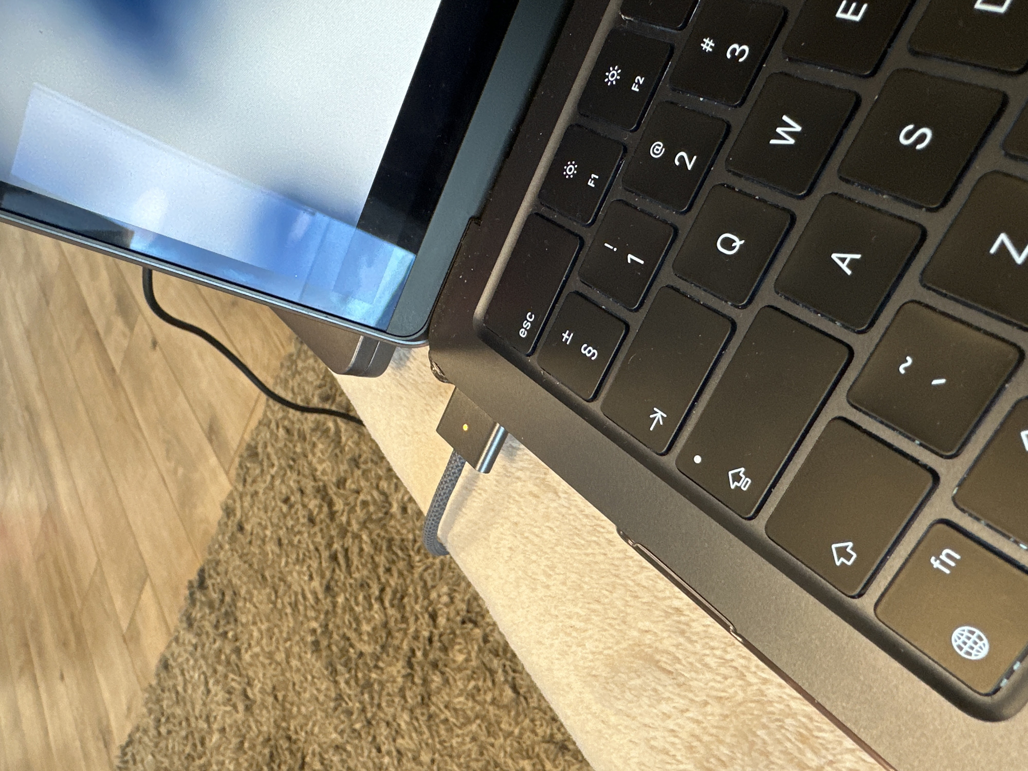 A close-up photograph depicts a portion of a dark gray laptop keyboard with keys labeled "esc," "F1," "F2," "1," "2," "3," "Q," "W," "E," "A," "S," "Z," "fn," and other symbols, next to a laptop screen reflecting the environment. A gray braided charging cable is connected to the laptop. The laptop rests on a light-colored surface, which appears to be a table or desk. A rug or carpet is visible at the bottom left of the image. The location is not specified, and the city of Sofia, Bulgaria, cannot be determined from the image.