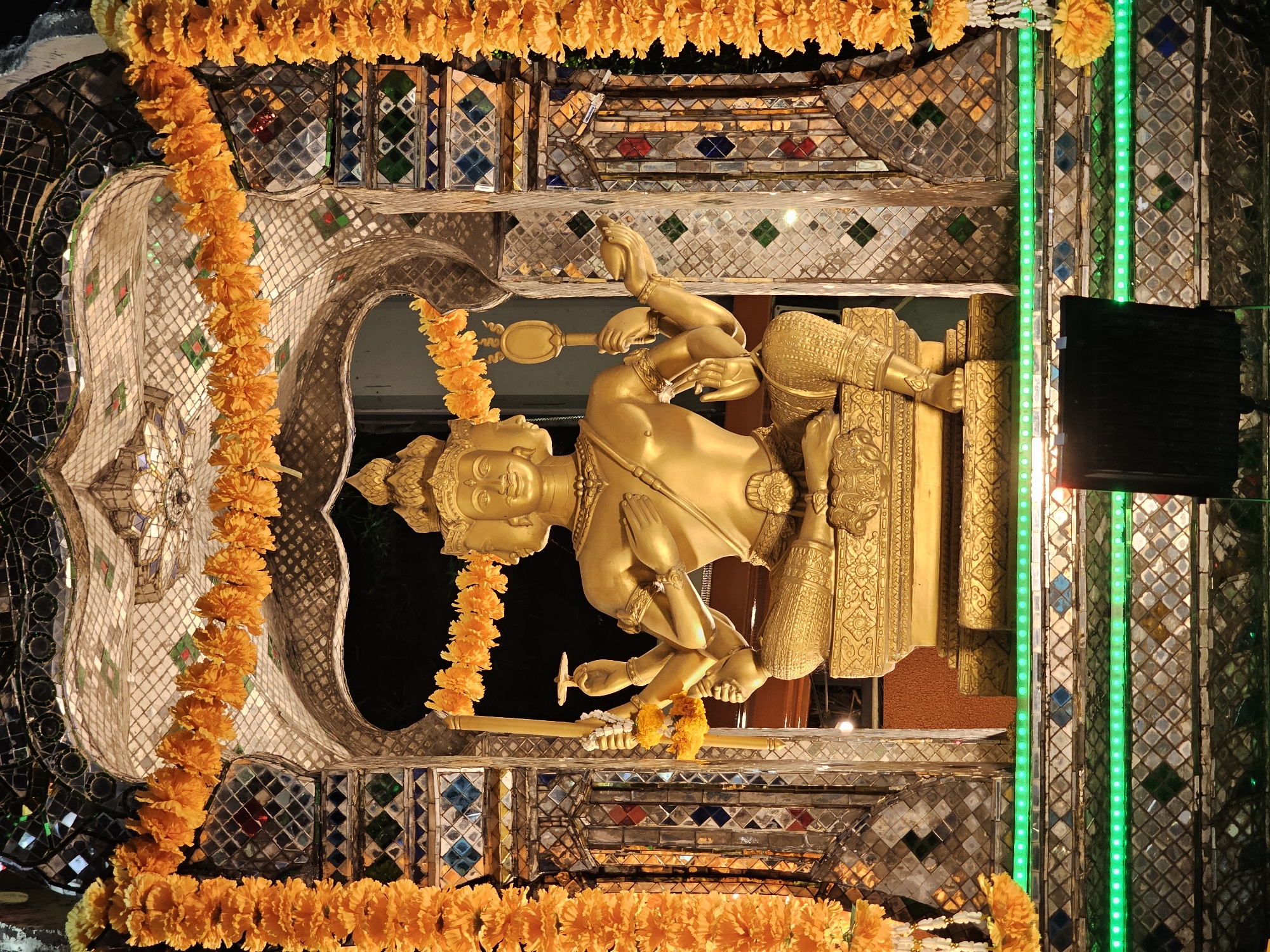 The image shows a golden statue of a four-faced deity, likely Brahma, seated in a shrine decorated with orange marigold garlands and colorful mirrored tiles. The shrine is illuminated by green LED lights at its base. The statue is depicted with multiple arms, some holding symbolic objects. The overall scene suggests a religious or cultural site in Bangkok, Thailand, possibly a temple or a shrine frequented by devotees. The lighting and decorations indicate a festive or devotional atmosphere.