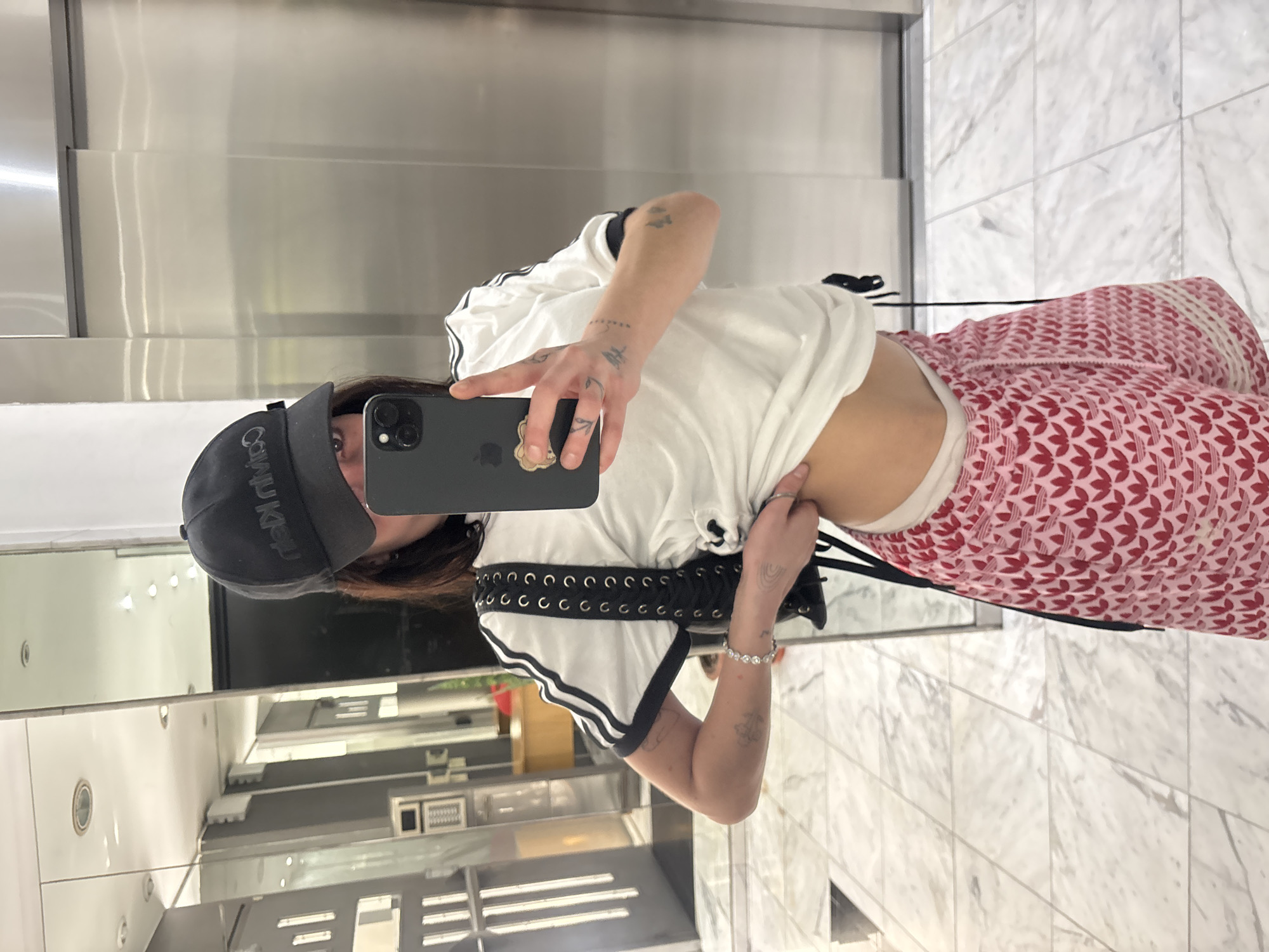 A person stands in what appears to be an elevator, taking a selfie with a black iPhone. The background features stainless steel elevator walls and marble flooring. Reflected in a mirror on the left is a retail space with shelves and racks. The person is wearing a black baseball cap, a white t-shirt with black stripes on the sleeves, a black corset-style belt, and pink patterned pants. The person's left arm is visible, adorned with tattoos and a bracelet. The image is taken in portrait mode and appears to be rotated 90 degrees clockwise. There is no evidence indicating the location is in Buenos Aires, Argentina.