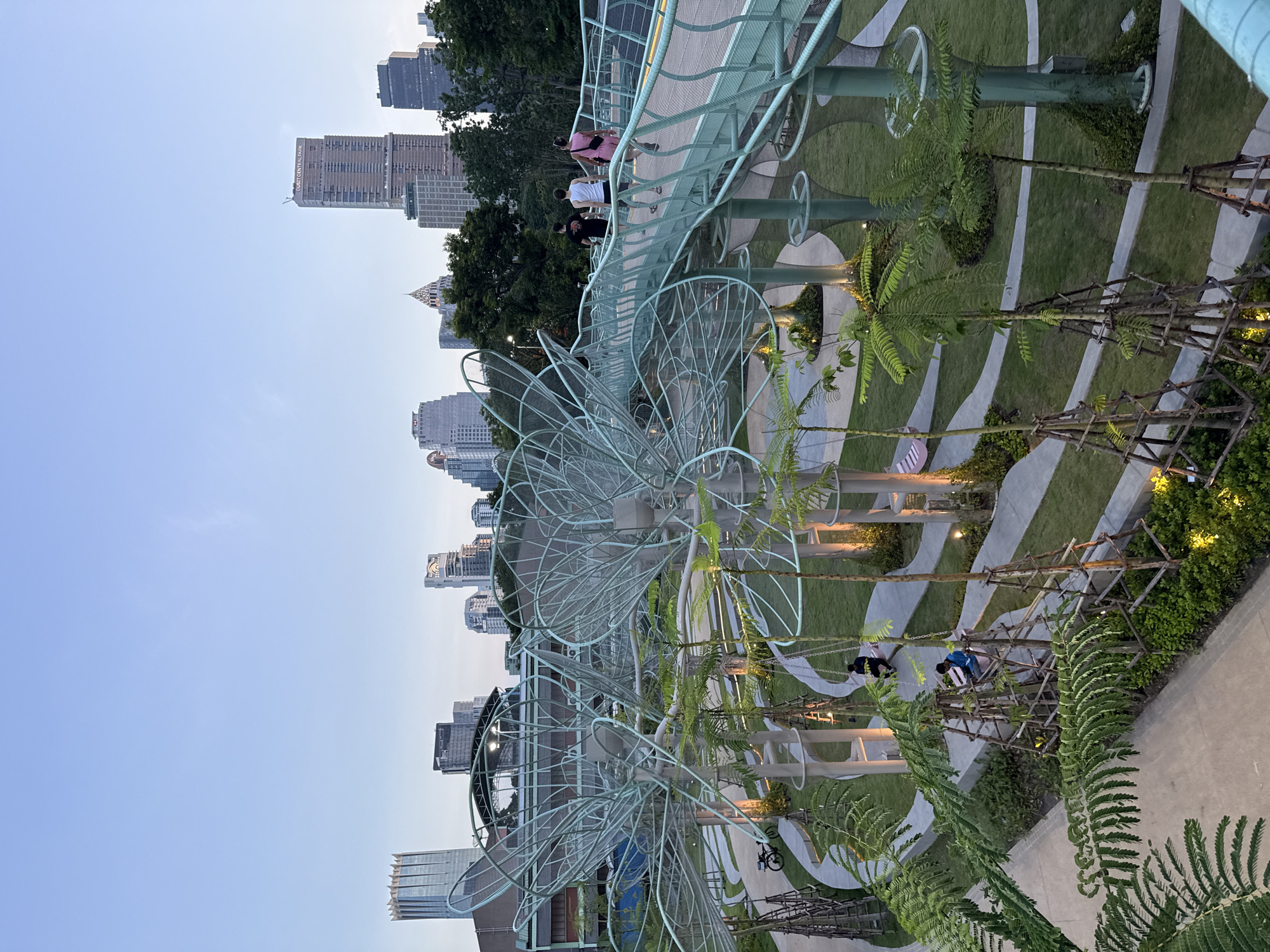 This is a high-angle, medium shot of a modern elevated walkway in a park in Bangkok, Thailand. The walkway, constructed of light green metal latticework, curves through the scene, leading the eye towards a distant cityscape of tall skyscrapers. Three people, two men and one woman, are walking away from the camera on the walkway. Below the walkway, a lush green park features winding pathways, manicured grass, and young trees supported by wooden scaffolding. Several people are seated on swings and benches in the park. The sky is a clear, pale blue, indicating either dawn or dusk. The overall scene depicts a contemporary urban park designed for recreation and relaxation amidst the city's dense architecture.