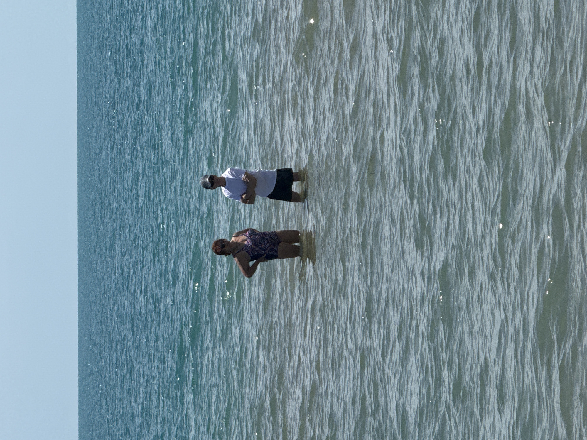 Two individuals stand in the sea, with the horizon visible in the distance under a clear, pale blue sky. The person on the left is a woman wearing a dark floral swimsuit. She has her hands on her hips and is looking towards the right. To her right, a man stands with his arms crossed. He is wearing a white t-shirt and dark shorts and a grey baseball cap. Both are submerged in the water up to their mid-thighs. The sea is a deep blue-green with gentle ripples on its surface, reflecting sunlight. This scene is located on the coast of Nesebar, Bulgaria.