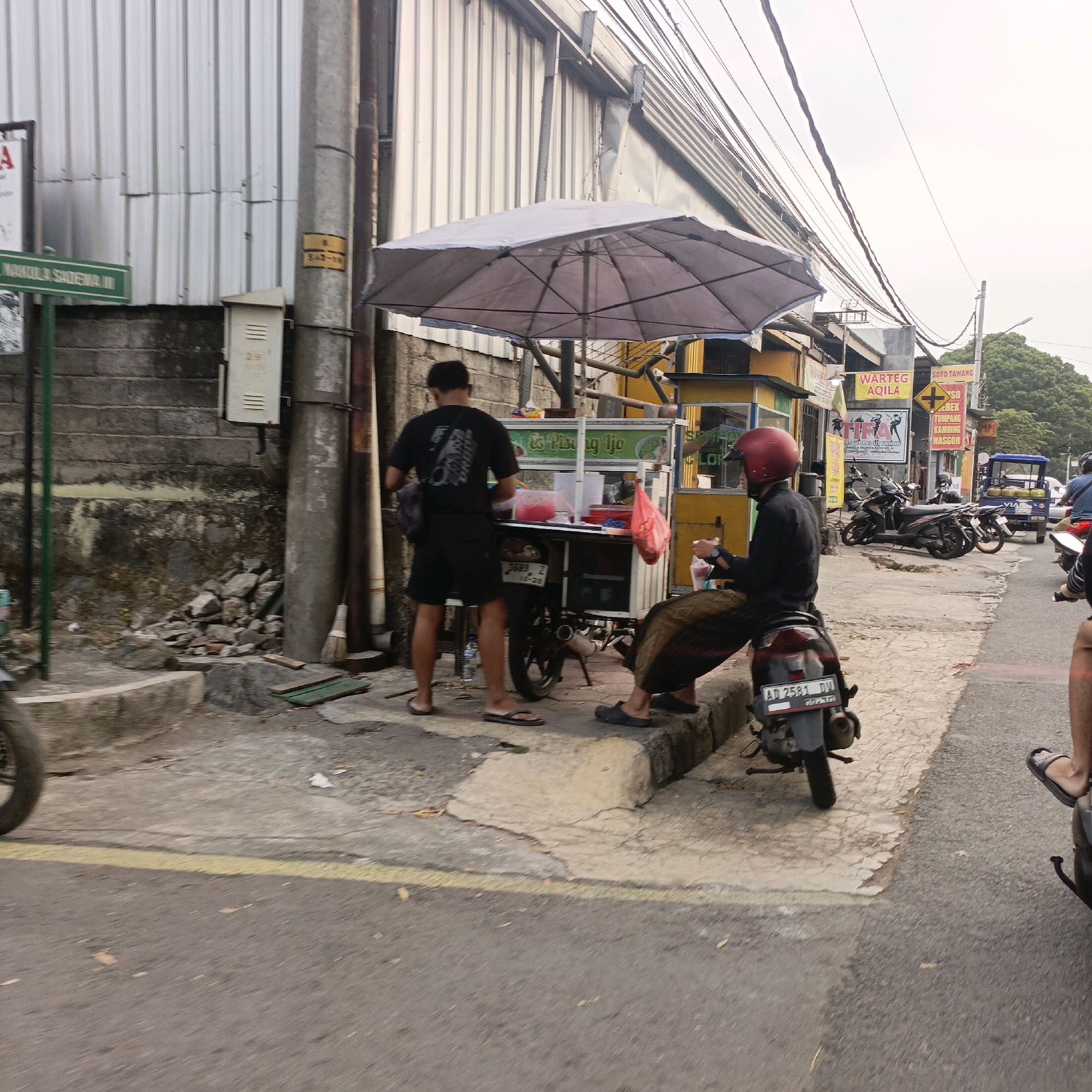 The image depicts a street scene in Salatiga, Indonesia. In the foreground, a person is seated on a motorcycle with a red helmet. Beside them, a food cart with a gray umbrella is set up. A man in a black t-shirt and shorts stands by the food cart, attending to it. A variety of motorcycles are parked along the street. In the background, there are buildings with metal siding, electrical wires, and street signs. One sign reads "WARTEG AQILA" and another indicates "SOTO TAWANG". The environment appears to be an urban street with some debris on the ground.