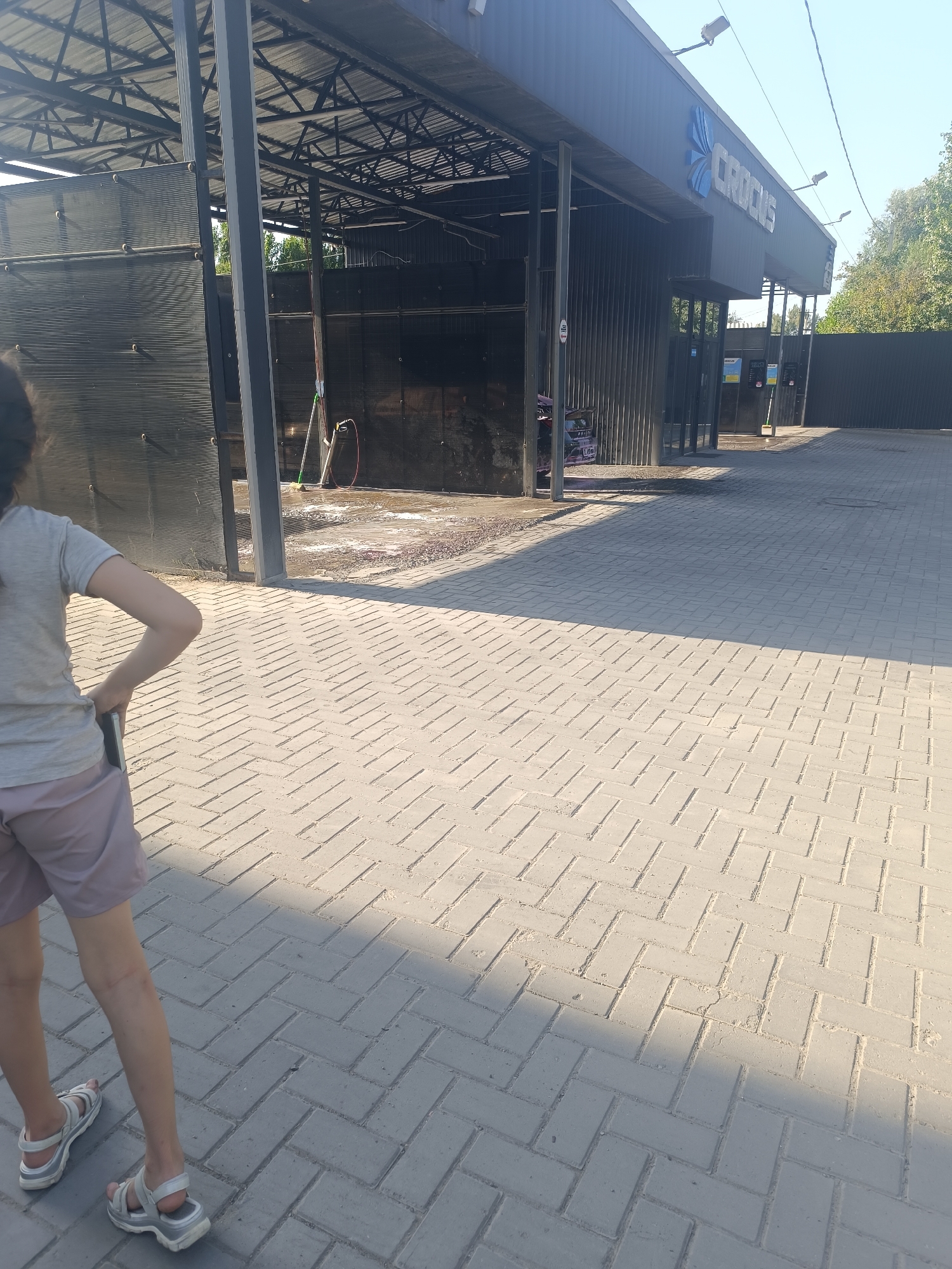 A person is standing on a brick-paved area outside a car wash station. The car wash station has a metal roof structure and dark corrugated metal walls. The "CROCUS" logo is visible on the front of the building. The ground is wet, indicating recent use of the car wash. There are various cleaning tools and equipment visible near the car wash bays. The scene is outdoors, with trees and a fence in the background. The sky is clear and blue. The person is facing away from the camera, wearing a grey t-shirt and light-colored shorts, and appears to be holding a mobile phone.