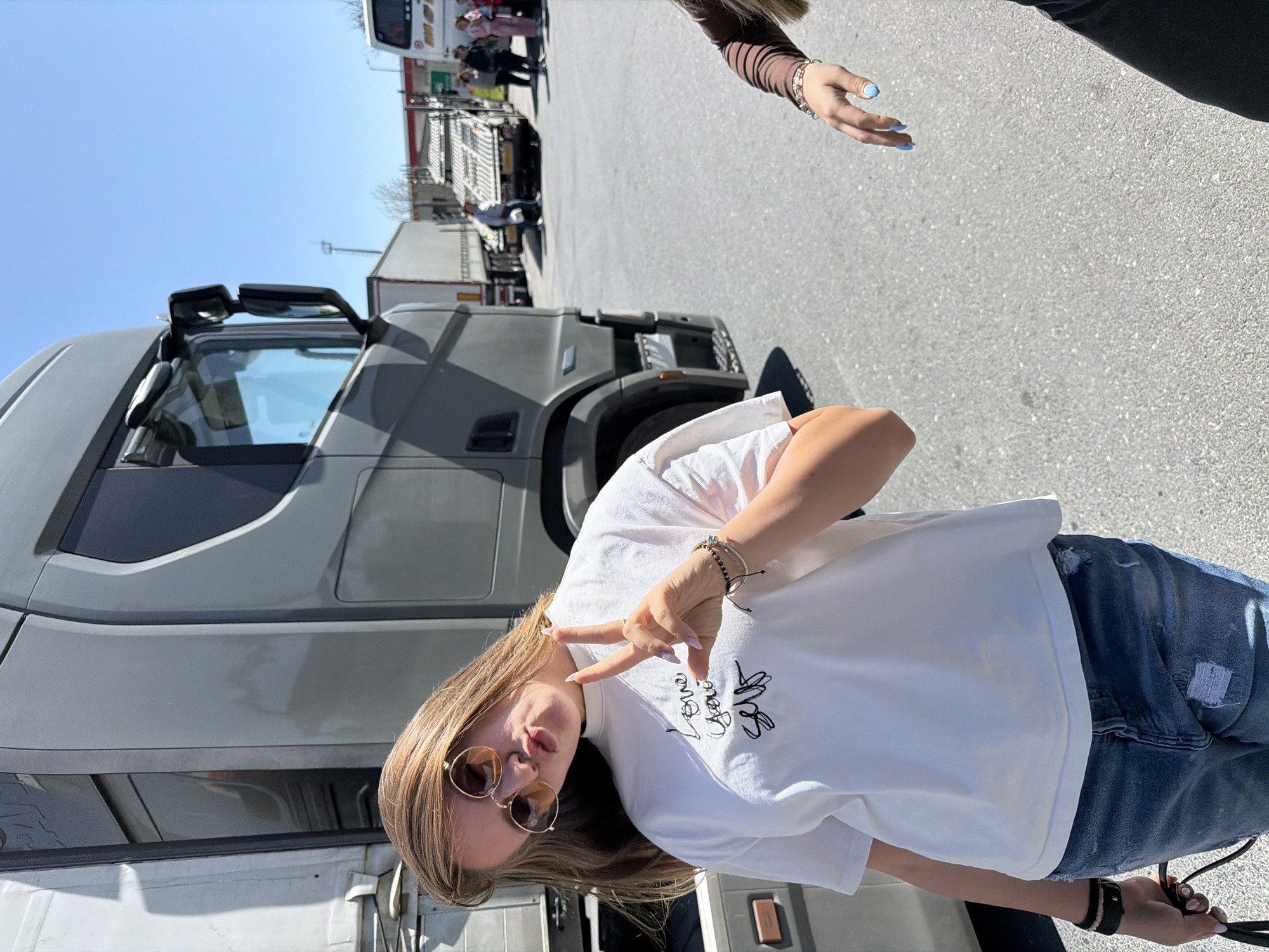 This media file captures a playful moment in a truck stop or large parking area in Çekmeköy, Turkey, during bright daylight hours.
The primary subject is a young woman with long, light brown hair, wearing round sunglasses, a loose white t-shirt, and blue ripped jeans. She is striking a pose, making a peace sign with her right hand while pouting her lips. On her left wrist, she wears a black watch and several bracelets, and she holds a black strap or bag in her left hand.
A prominent grey semi-truck occupies the left side of the frame. In the background, an expansive asphalt lot extends, revealing more trucks, trailers, and a white bus. Several other people are visible near the bus in the distance. Partially visible in the top right is another person's arm and hand, featuring light blue nail polish and a bracelet.
The clear blue sky and strong shadows indicate a sunny day. On the front of the woman's white t-shirt, black embroidered text is visible, which clearly spells "Love You" in a cursive style, surrounded by other subtle graphic elements. The overall scene suggests a casual stop at a busy transport hub.
