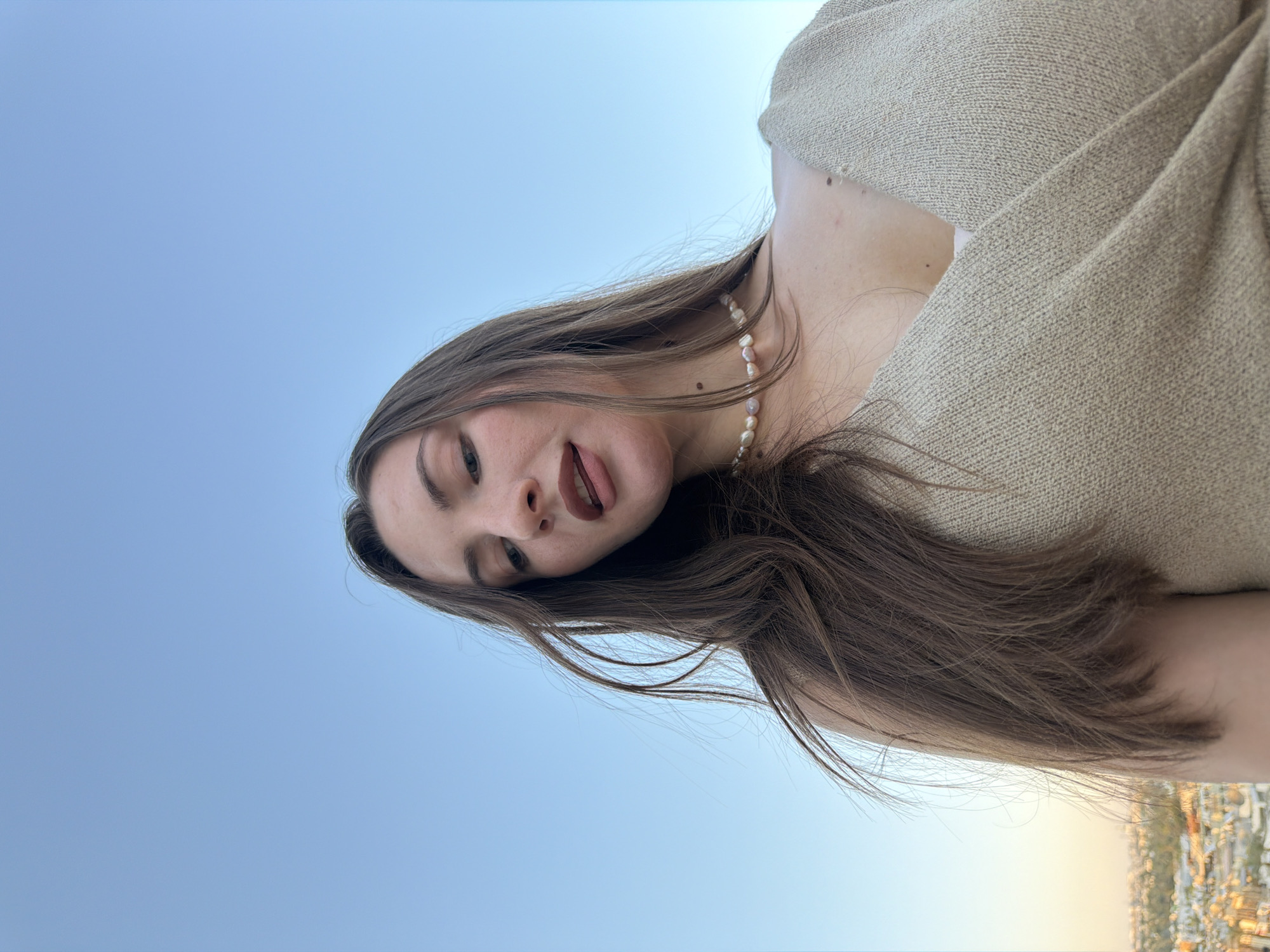 A close-up, low-angle, vertical shot captures a young woman with long, brown hair looking up and sticking her tongue out at the camera. She is wearing a light-colored, textured sweater and a pearl necklace. Her facial expression is playful and mischievous. The background is a clear, pale blue sky. Below the sky, a blurred, hazy view of a city with buildings and distant lights suggests an urban setting, possibly at dusk or dawn due to the warm, diffused light. The location context is Sharm El Sheikh, Egypt. There is no visible text.